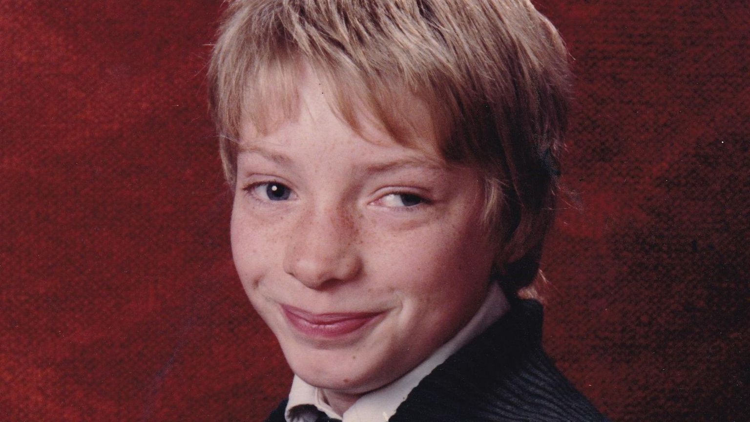 A young boy with sandy-coloured hair faces the camera, with a red background