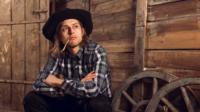 Tom Skelton sitting down with a piece of straw hanging out of his mouth, with hat on.
