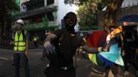 A protester masked and clothed in black is holding a flaming petrol bomb, arm back and ready to throw