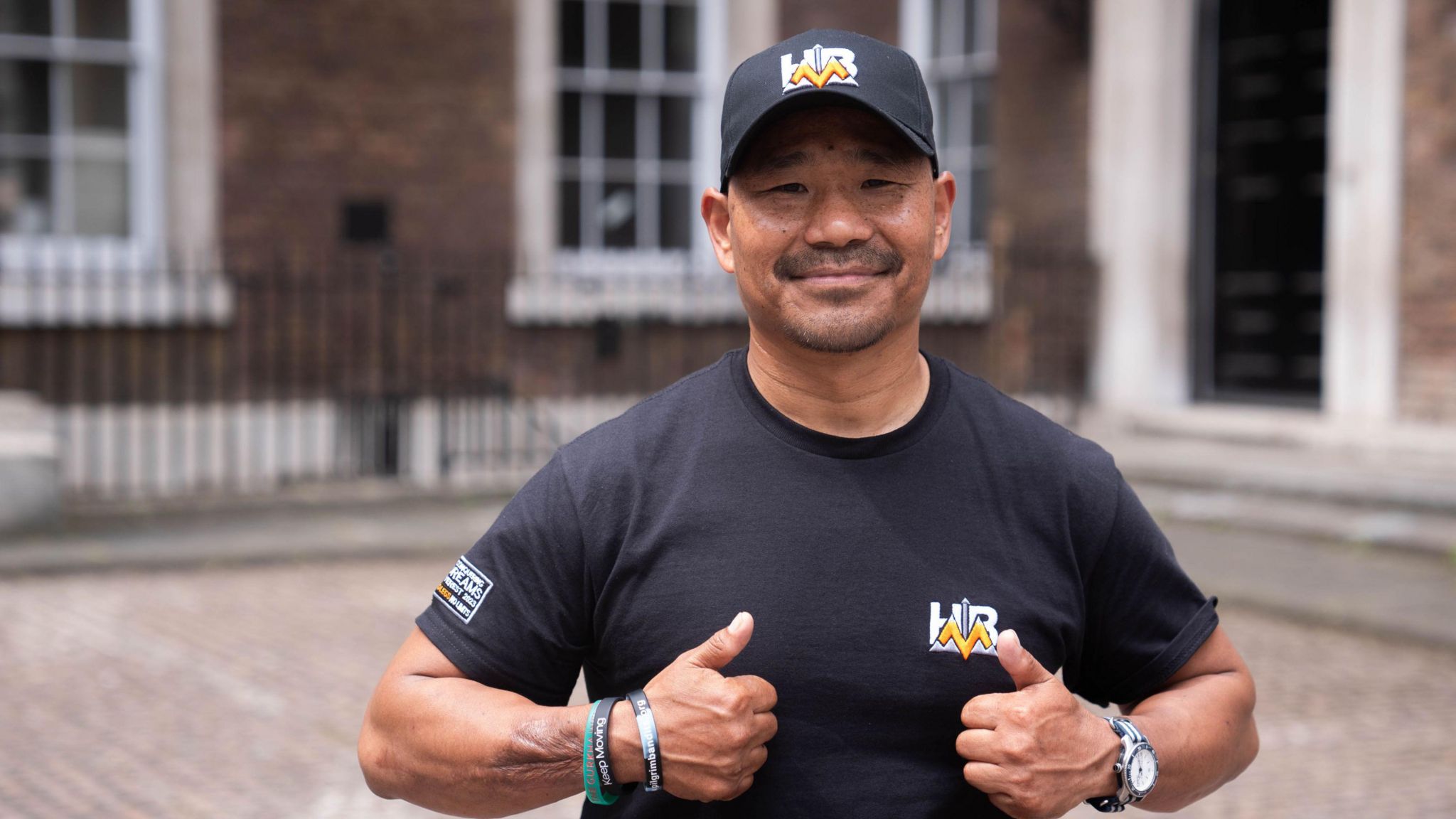 A man wearing a black hat and t-shirt, smiling with his thumbs up