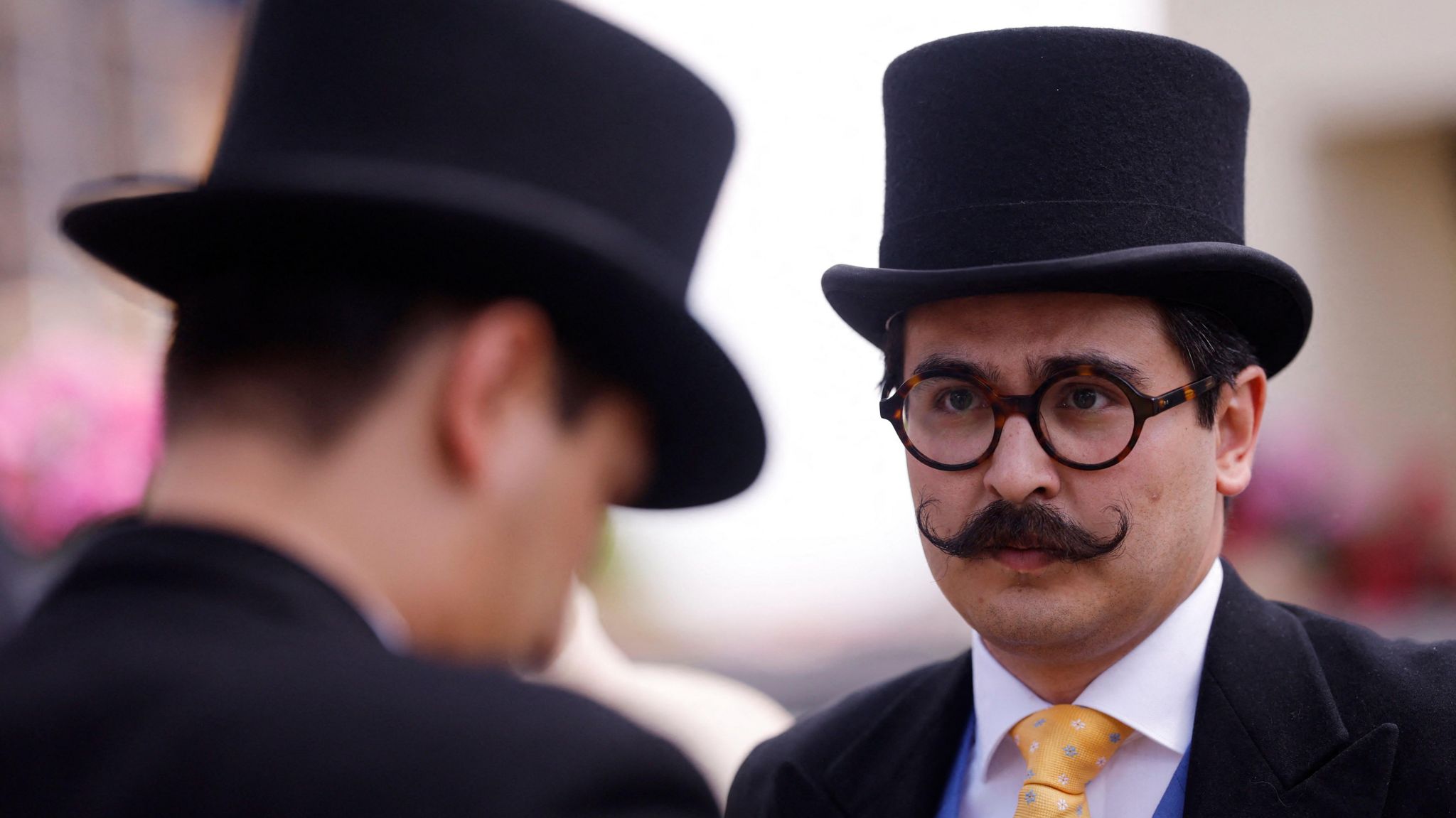 Two men wearing top hats, one facing the camera wearing round thick-framed glasses and a curled moustache