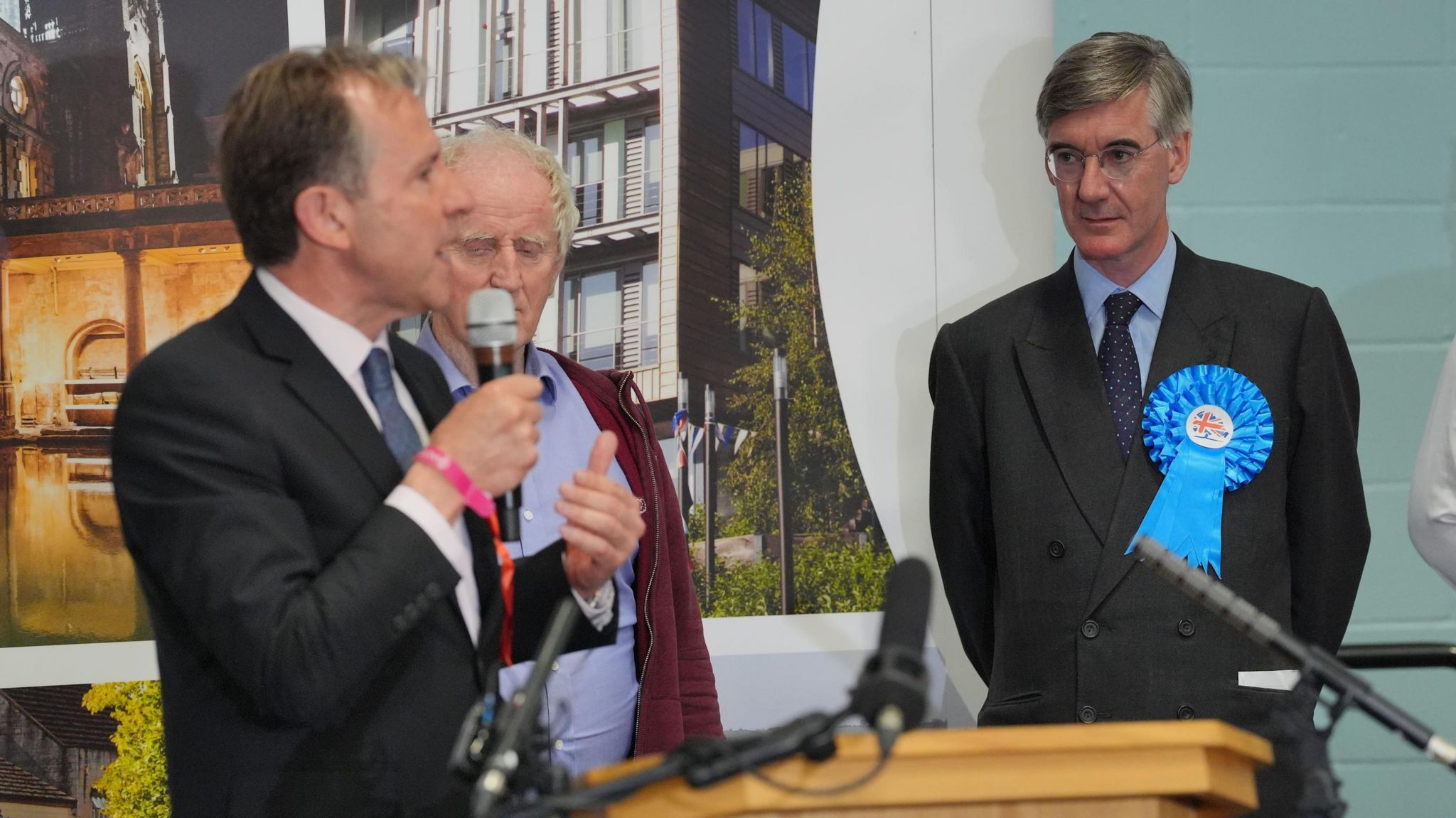 Jacob Rees Mogg wearing a blue rosette pinned to his suit looking at Dan Norris who is holding a microphone in front of a podium during the general election count result in July 2024