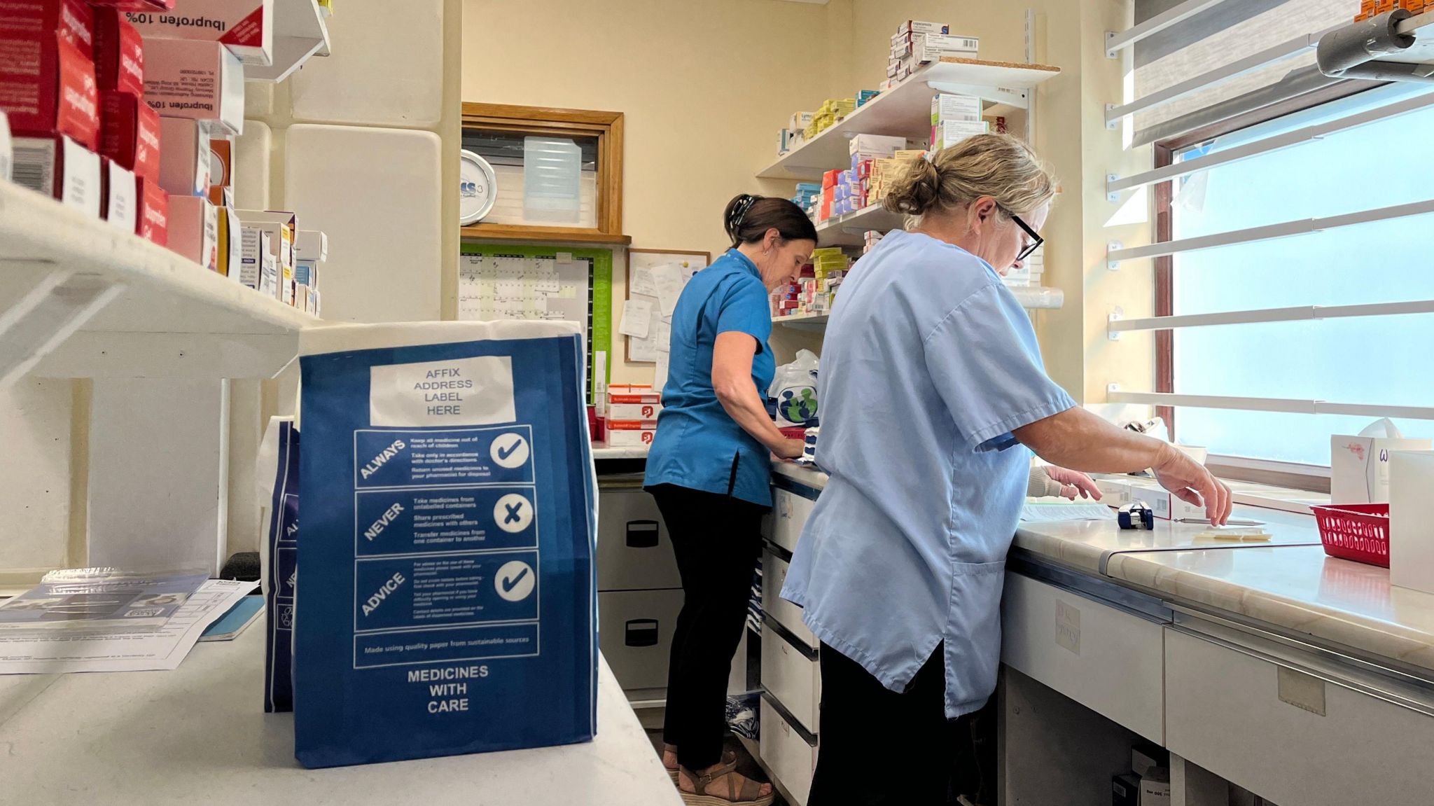 Two pharmacy staff at work in the dispensary. Shelves full of medication can be seen and bags of completed prescriptions stand on a counter.