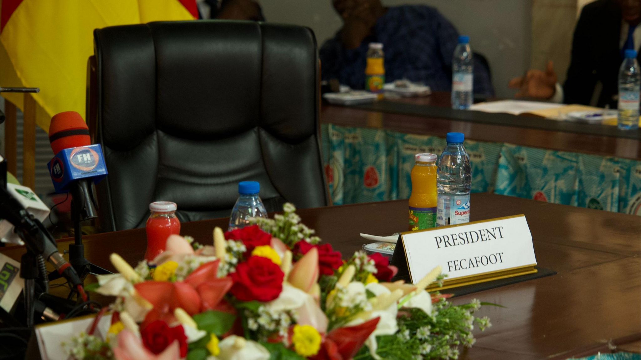 An empty chair sits behind a desk with a place card for the Fecafoot president, microphones and flowers