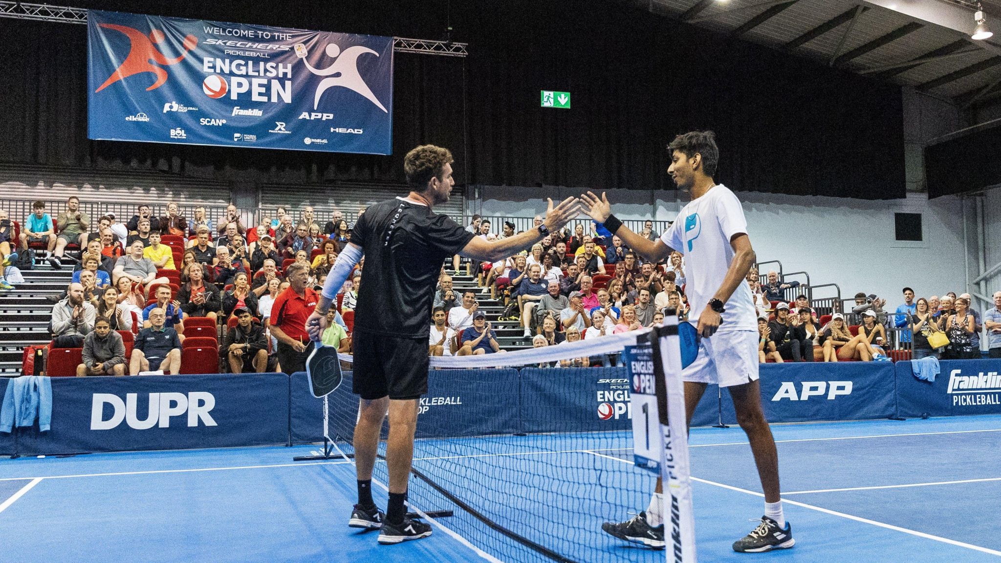 Two men shake hands over the top of a net. They are holding pickleball paddles. They are in a court with a blue floor, surrounded by blue banners. On the wall behind them is a banner that reads "Welcome to the Pickleball English Open". There is a crowd of people watching them, seated in stands.