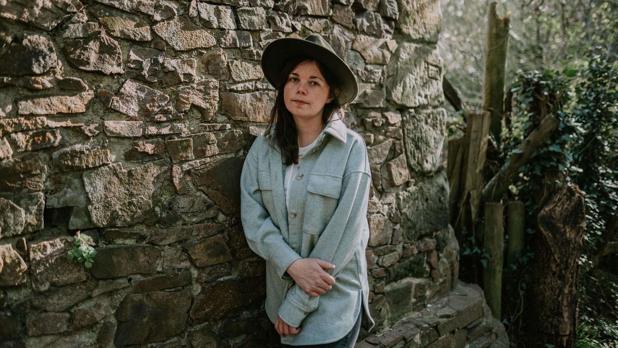 Jessie Reid in front of an old brick wall and she is wearing a blue coat and a wide-brimmed hat