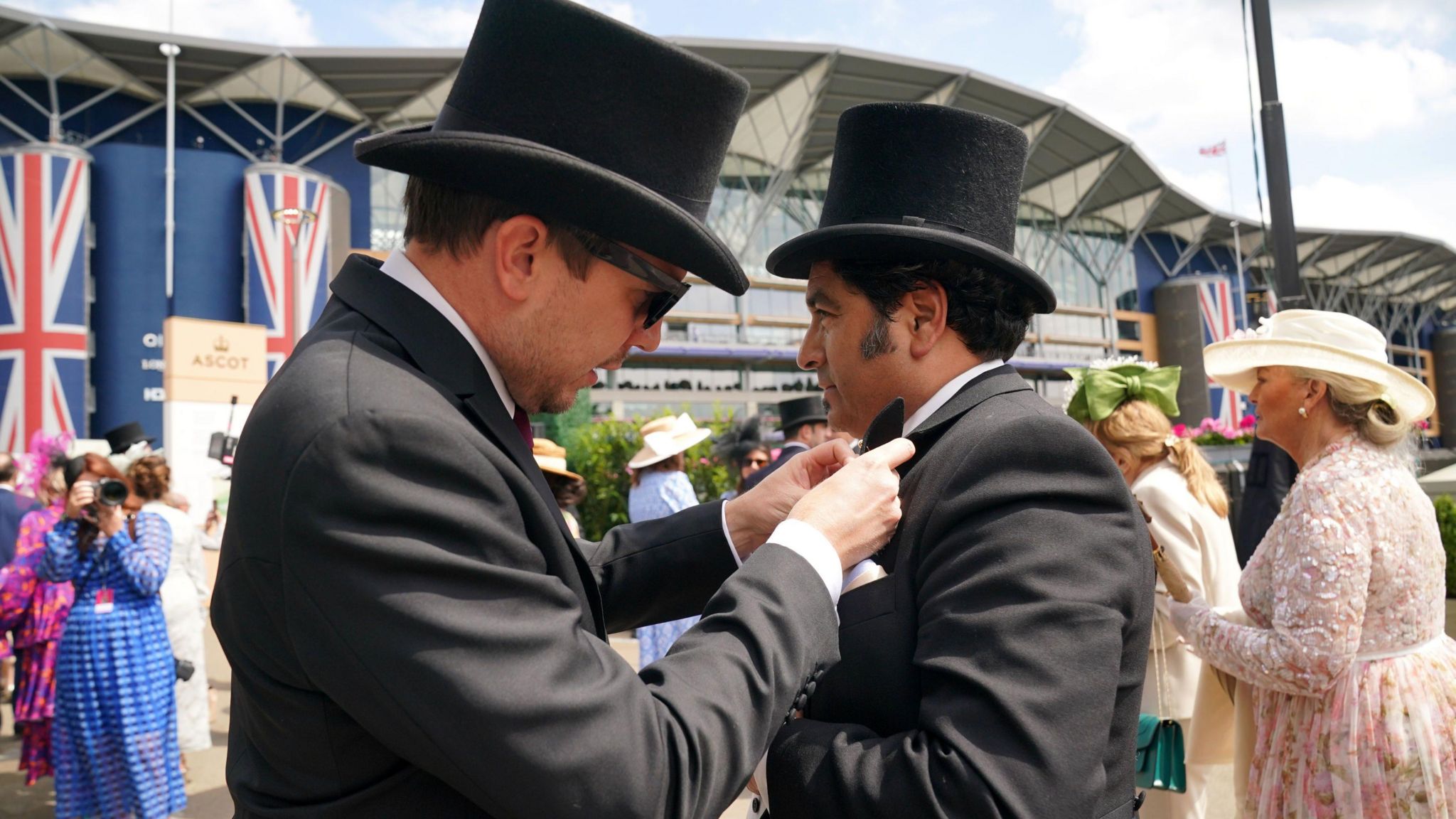 A man in a suit and top hat adjusts the pocket square of another man, with the Ascot Racecourse building in the background