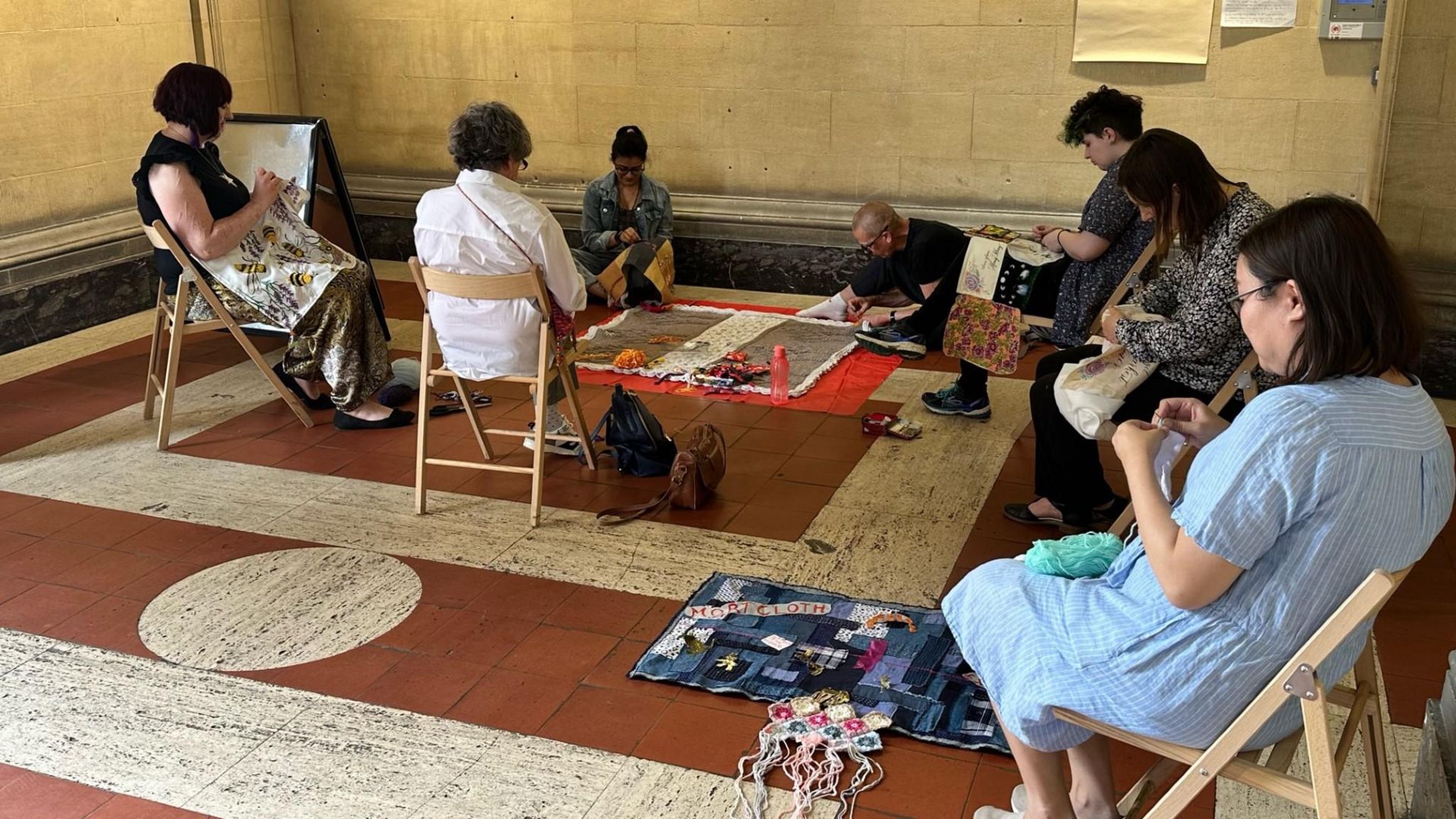 Sette donne sedute su sedie e sul pavimento che lavorano a pezzi di patchwork