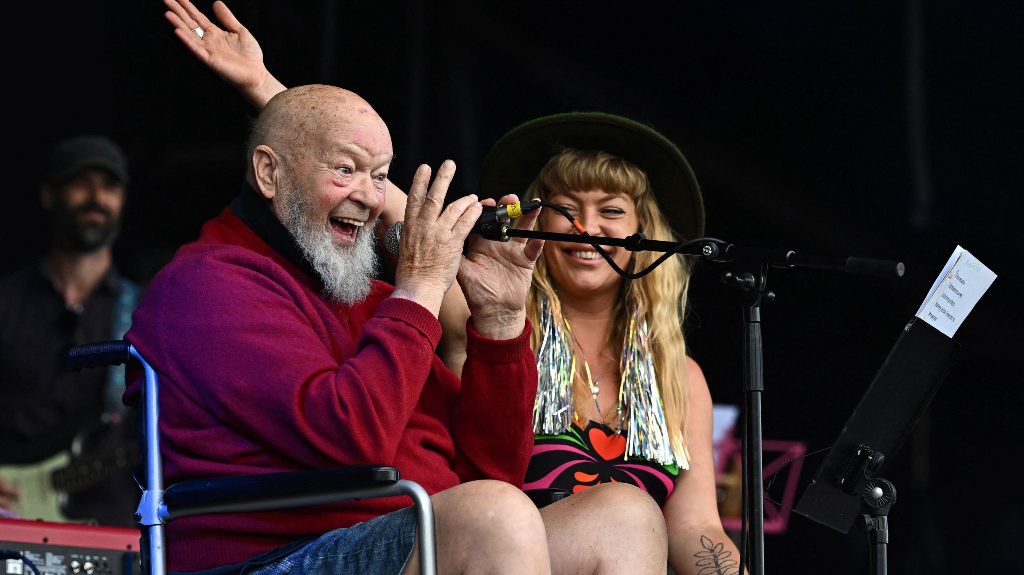 Image of Sir Michael Eavis on stage at Glastonbury. He is pictured wearing a red jumper. He is holding a microphone and smiling at the crowd. 