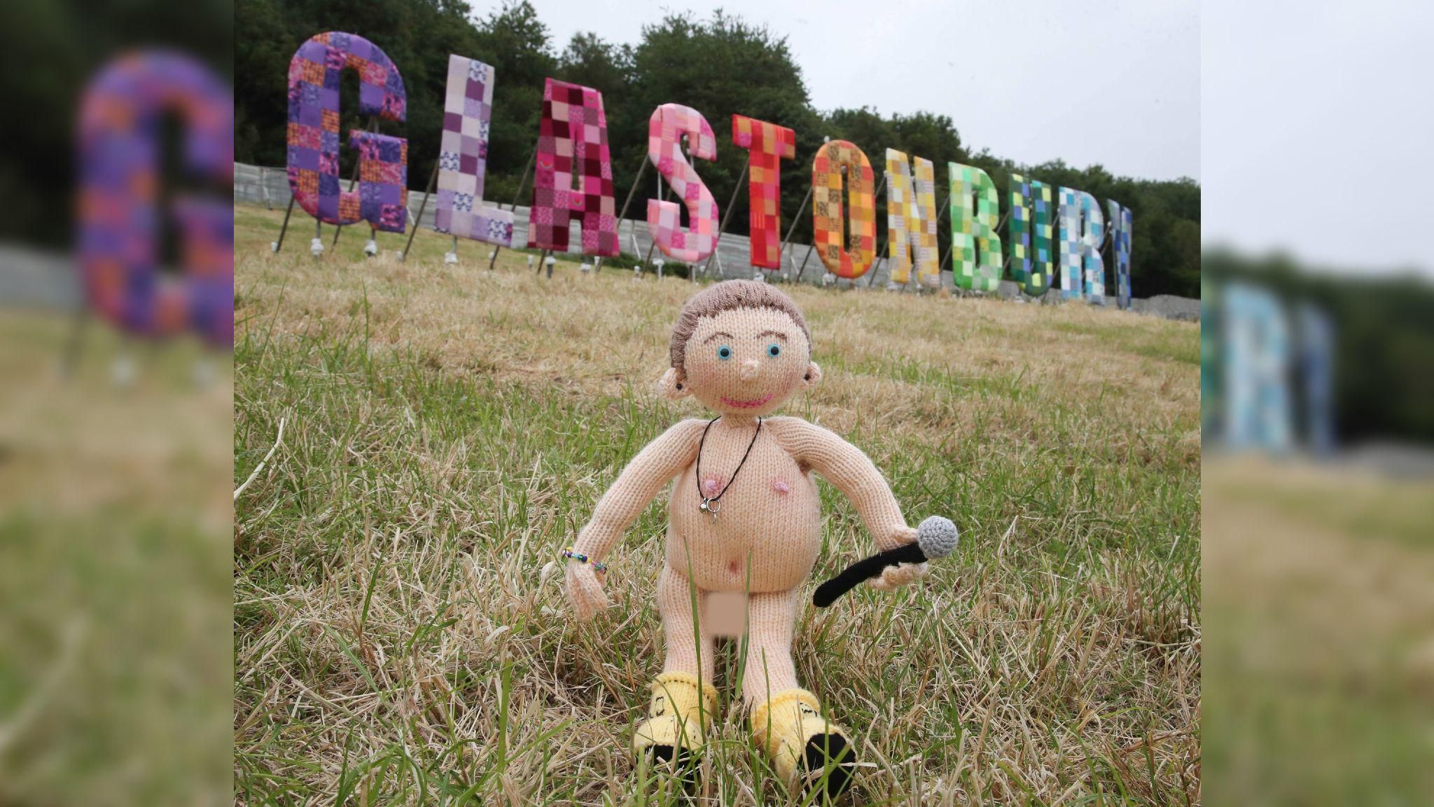 A knitted doll resembling Chris Martin standing on the grass in front of the colourful Glastonbury sign. He is naked and holding a microphone and wearing yellow boots
