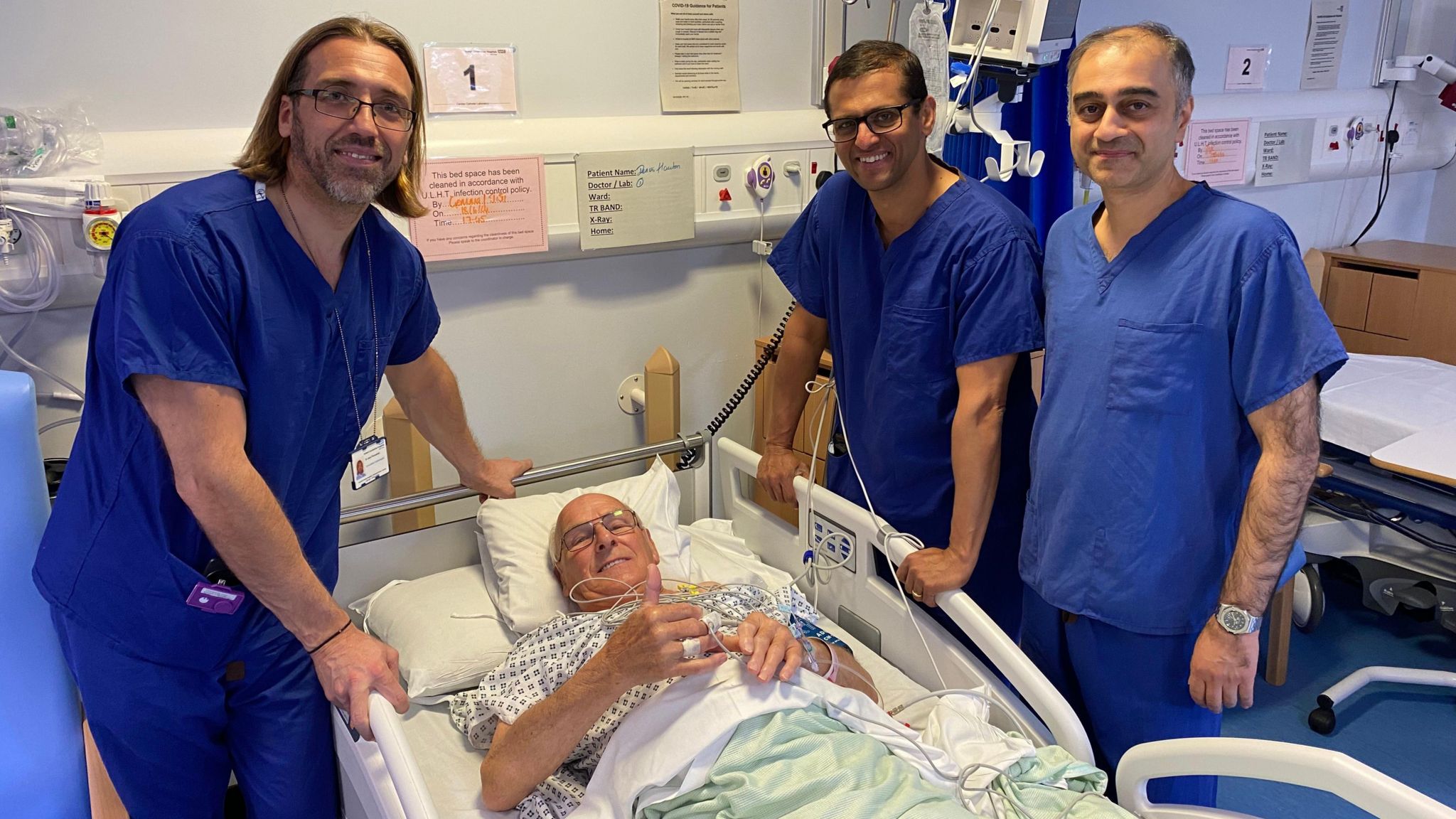 Hospital patient Dennis Hauton gives a thumbs up while laying in his hospital bed, surrounded by three doctors all wearing blue outfits