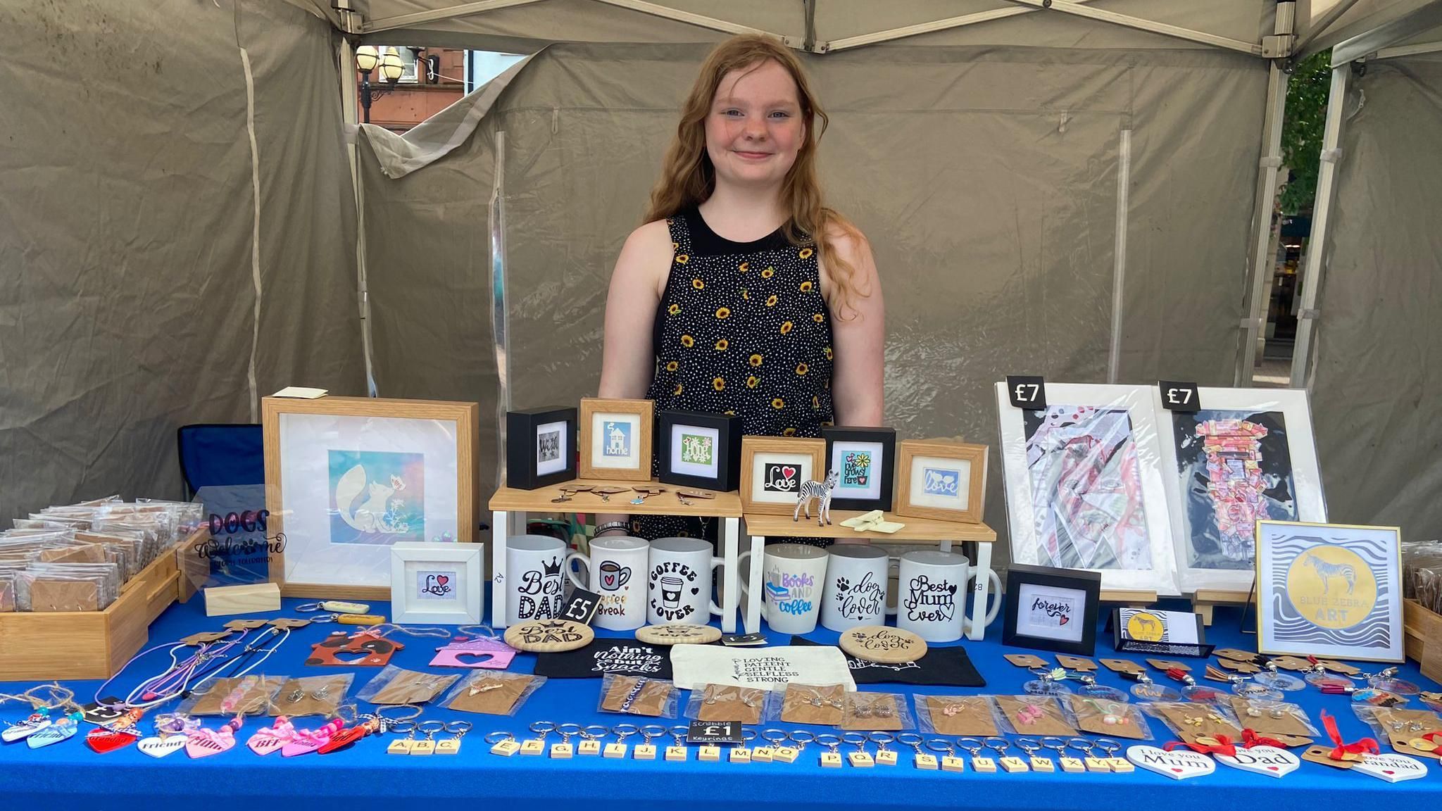 Teenager in flower dress stands behind stall filled with mugs, artwork and jewelry
