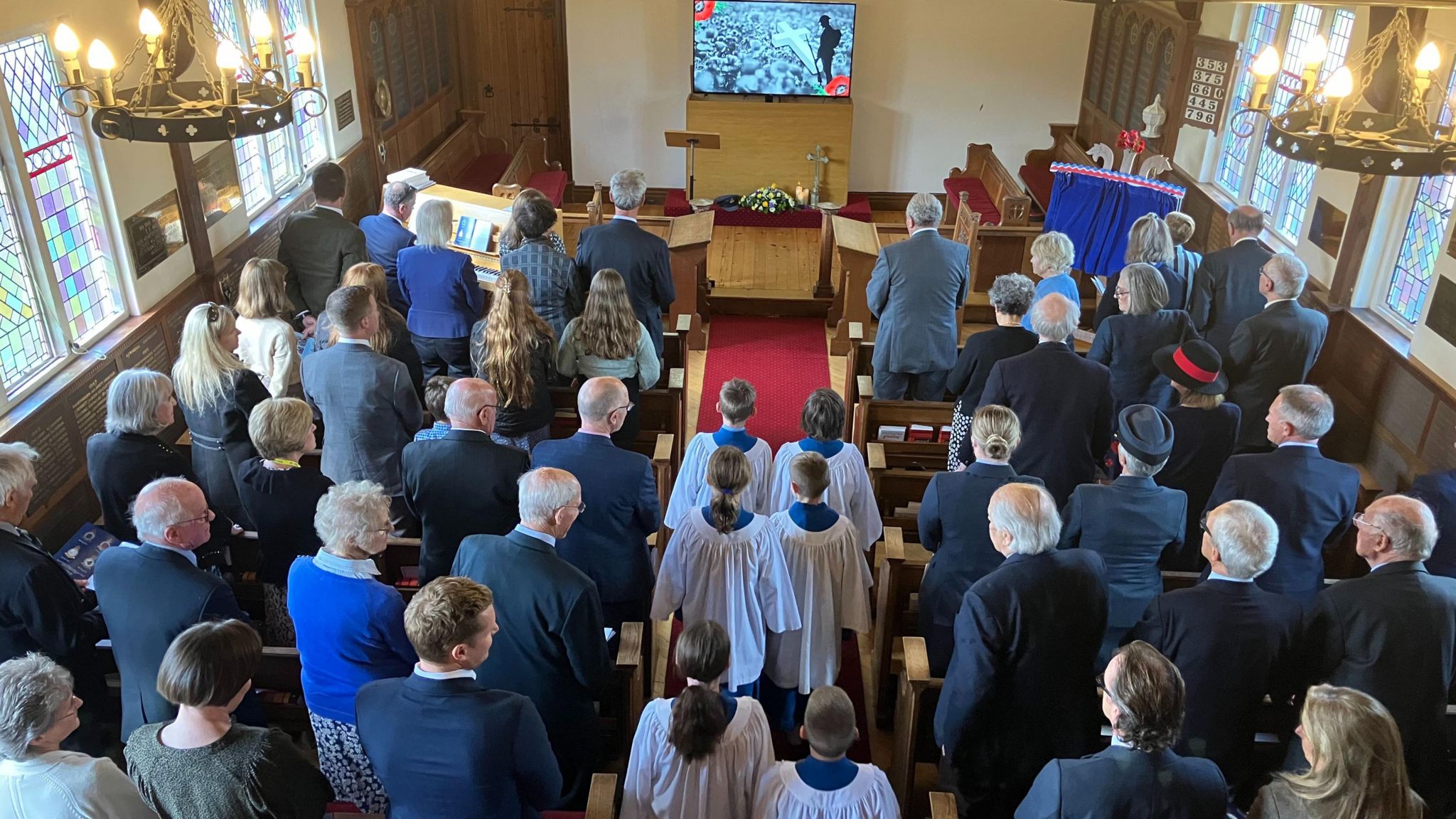 A photo of the ceremony take from above. It's a small chapel with stained glass windows on either side. All the seats are filled and a children's choir is walking down the aisle. At the front, the plaque is covered by a blue curtain.