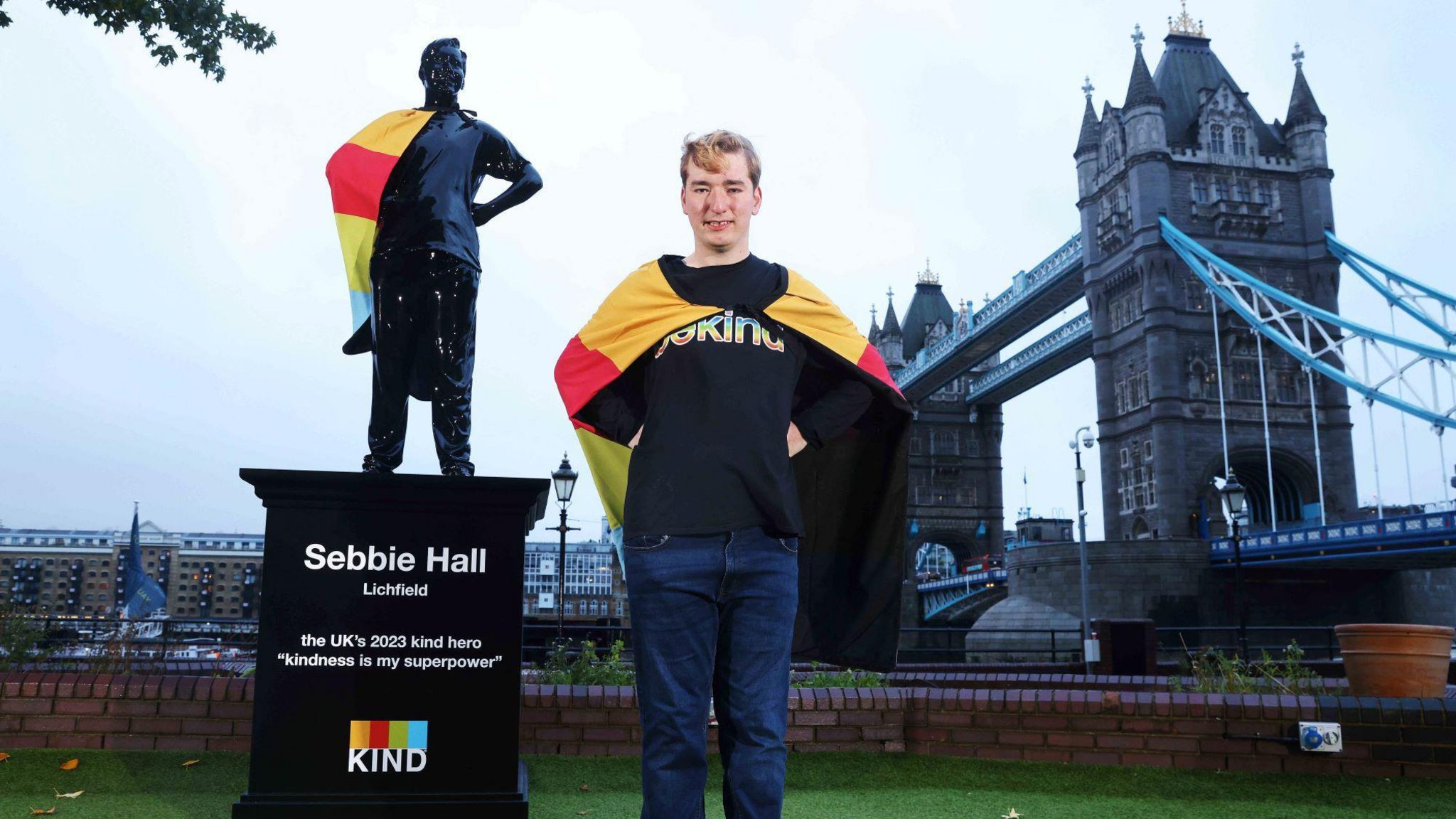 A man wearing a multicoloured cape stands next to a statue of himself, with London's Tower Bridge in the background