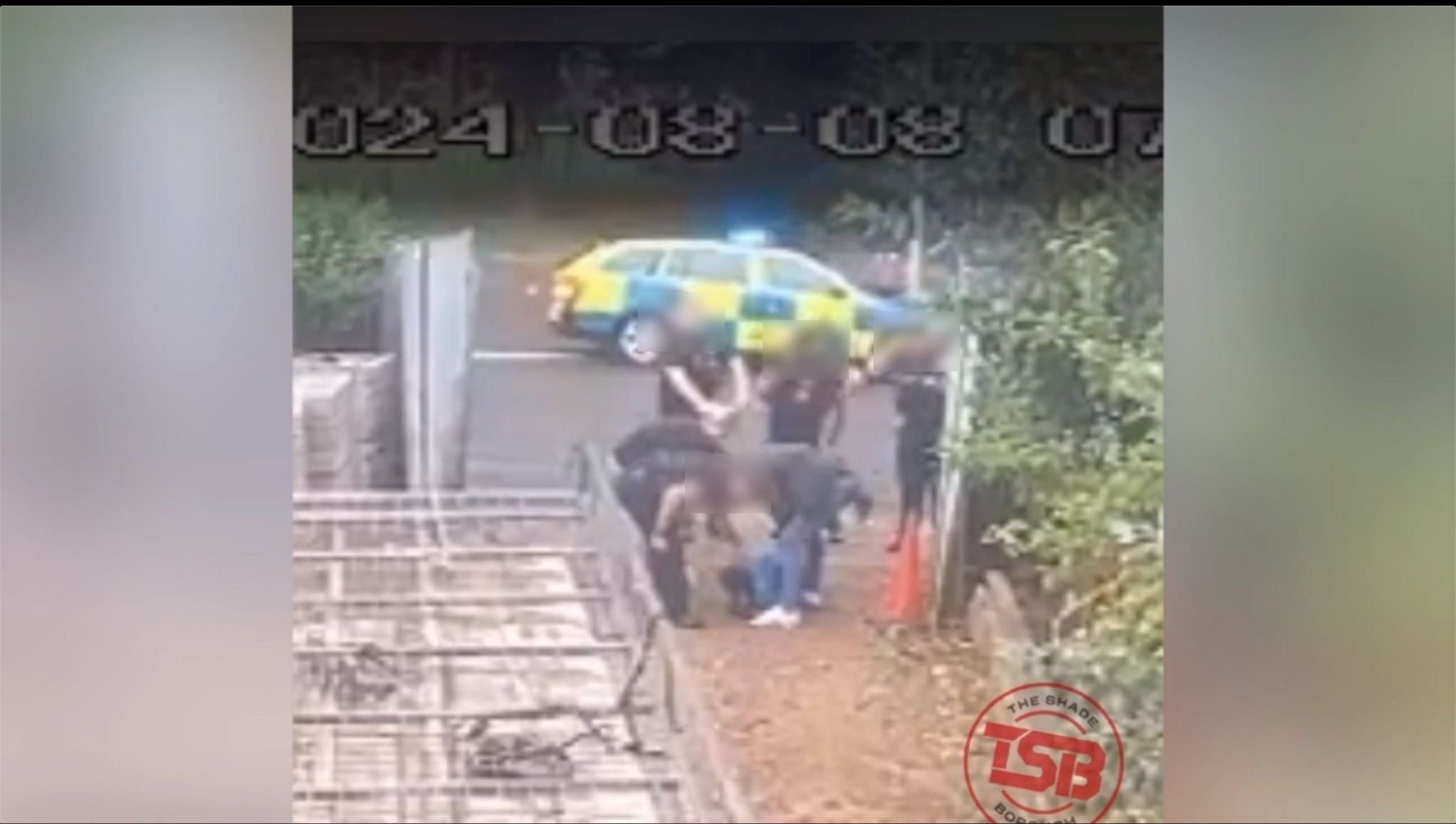 Five police officers crouch over a man wearing a blue jacket who is face down on the floor.  There is a wooden gate and hedge to the right and a police car is parked on the road behind