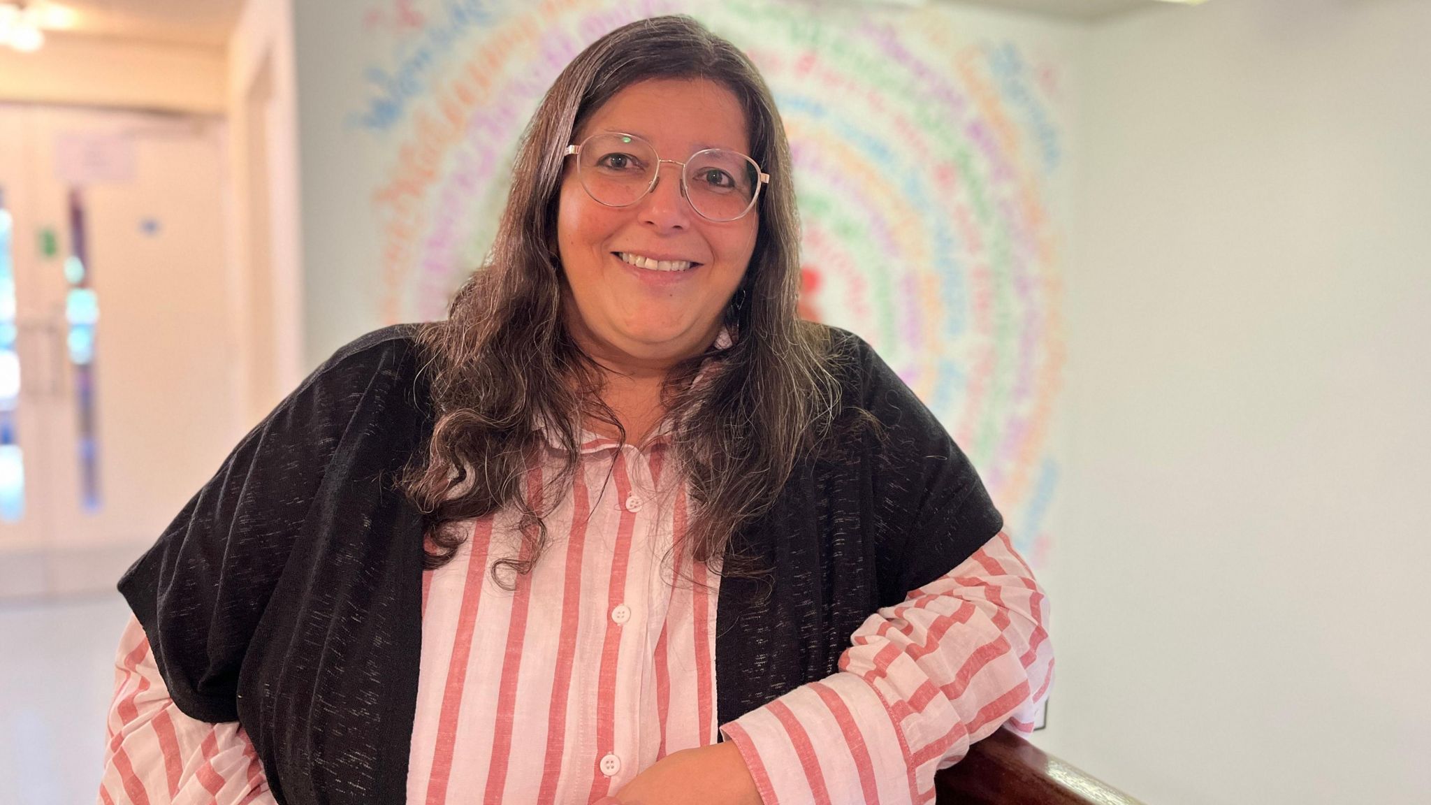 Maria Wilby wears a black shawl over a pink and white striped shirt, standing in front of an artwork designed by a refugee