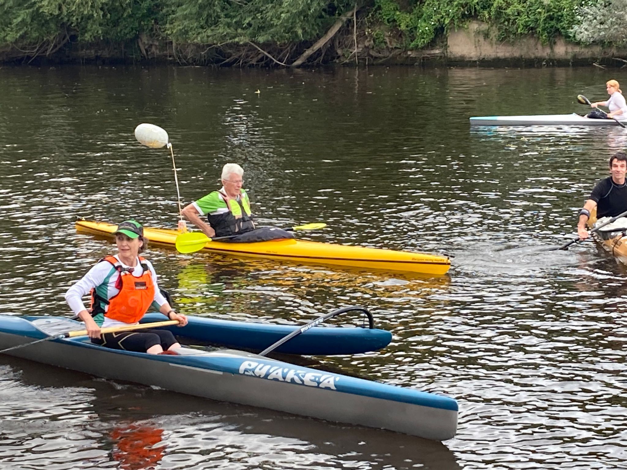 One woman wearing an orange lifejacket is paddling in a blue canoe and to her right a man in a yellow and black lifejacket is paddling in a yellow 