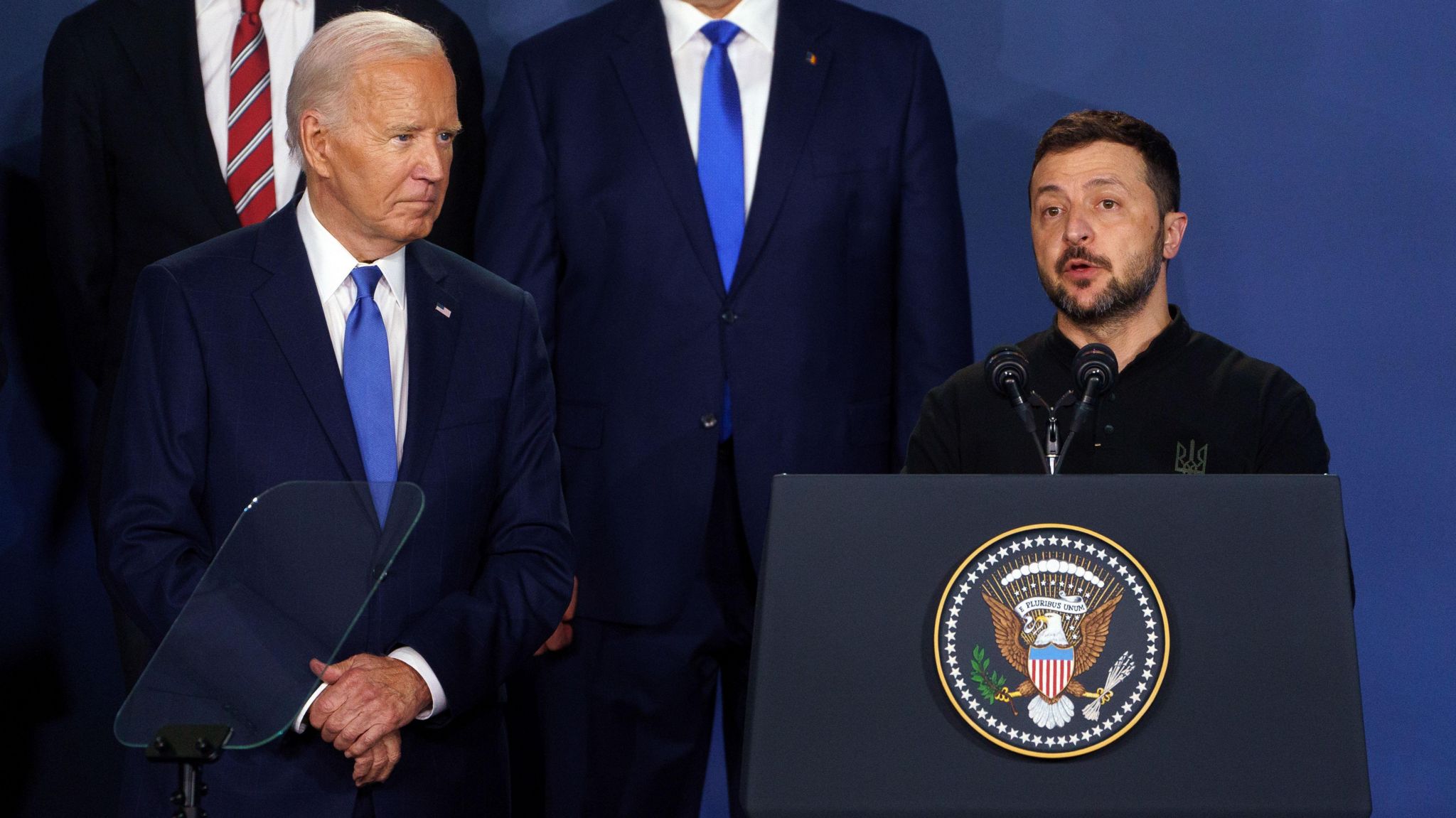 Wearing a blue tie and navy suit, US President Joe Biden stands next to Ukrainian President Volodymyr Zelensky who is speaking at a podium addressing the Nato Summit in Washington in July 2024