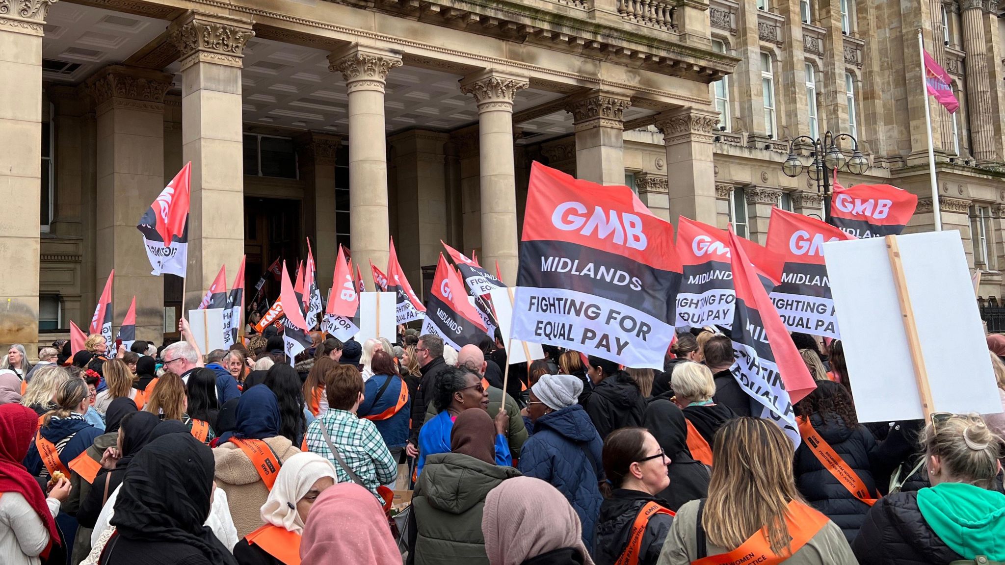 A crowd of strike supporters gather outside Birmingham City Council House waving flags displaying the message GMB Midlands Fighting For Equal Pay