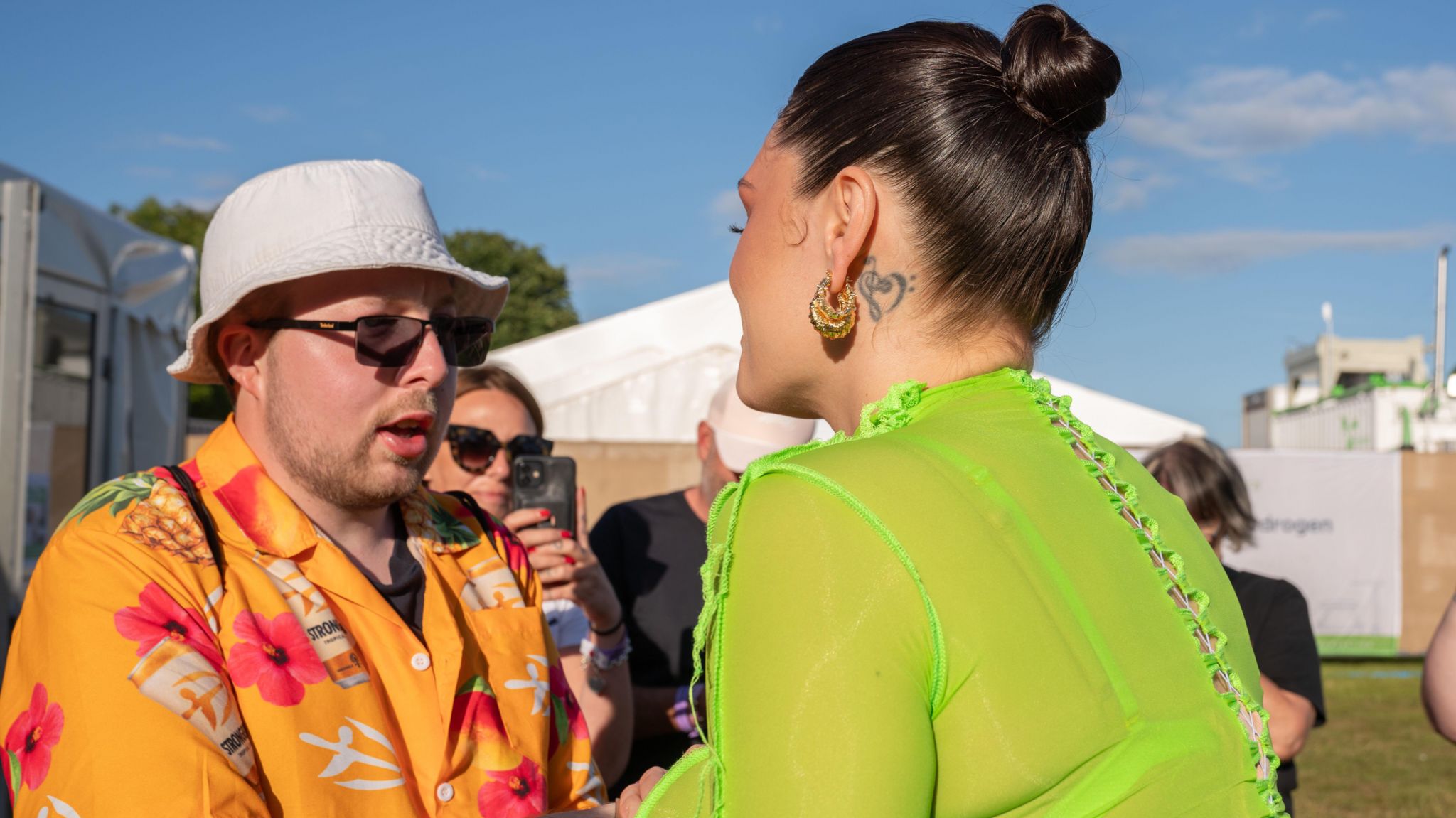Jessie J in a lime green outfit talking to Nathan who is wearing a orange shirt with pink flowers on. He is also wearing a white bucket hat and crowds of photographers and spectators are around watching the meeting