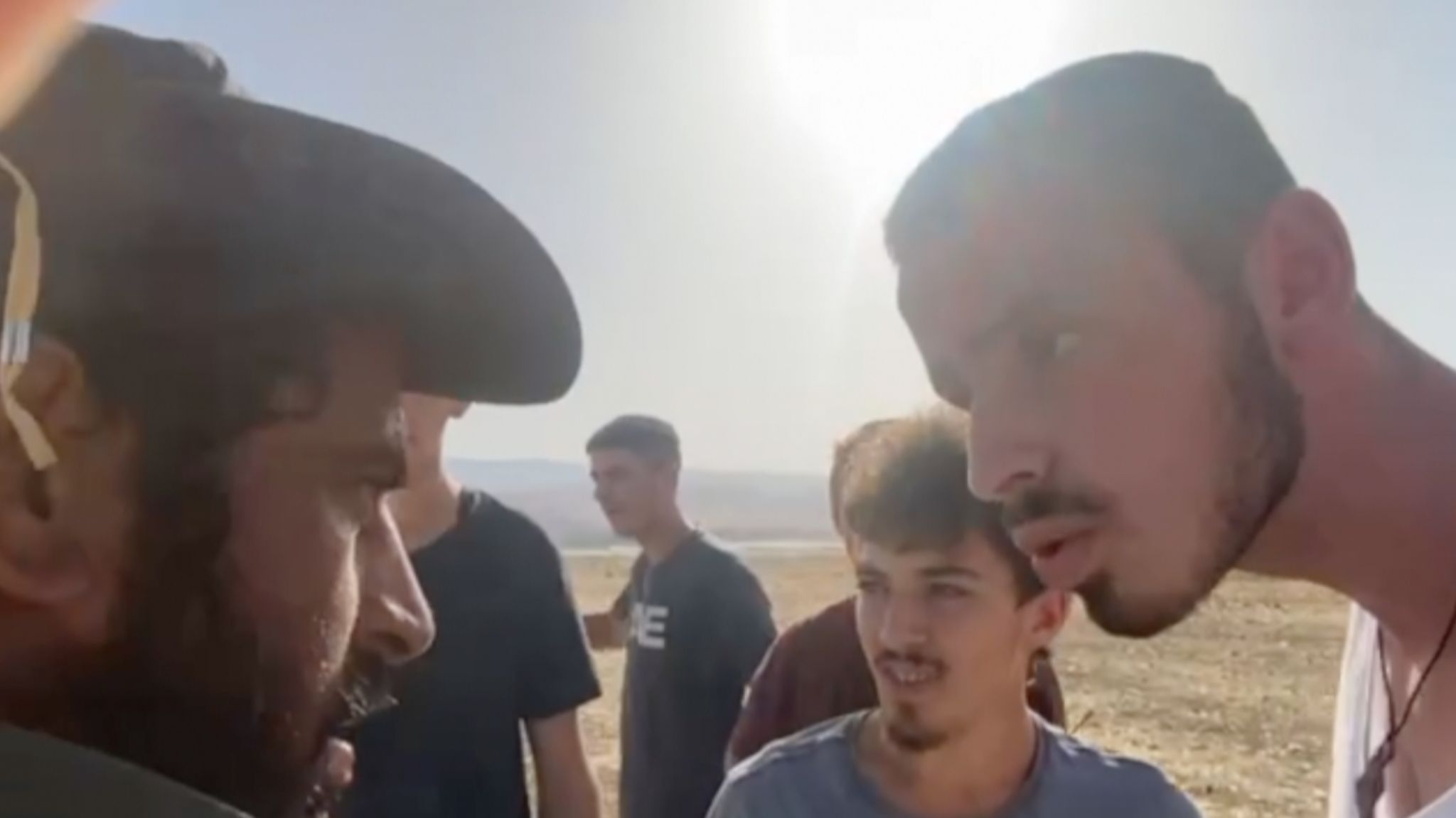 Ahmad, wearing a hat, appears to be harassed by settlers. It shows a close up side profile of Ahmed's face, with a man appearing to talk into his face. A number of other men are gathered around them.