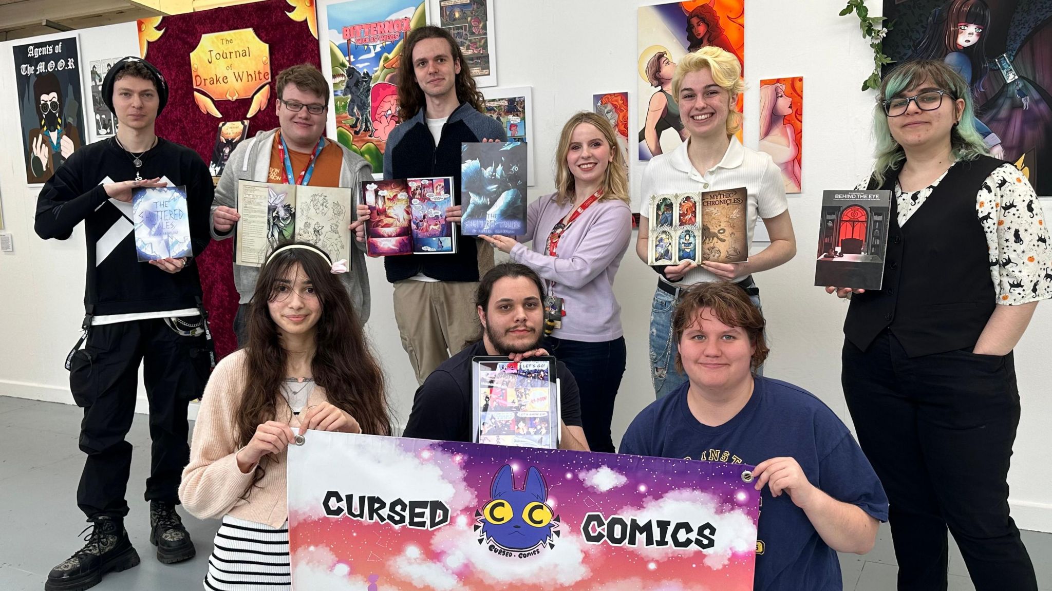 Nine comic book students stood in front of comic book designs that are pinned to a white wall. All are holding the comic books that they designed and illustrated. Two people are holding a pink and purple banner that reads "cursed comics" The banner has a picture of a blue rabbit with yellow eyes in the middle of it.