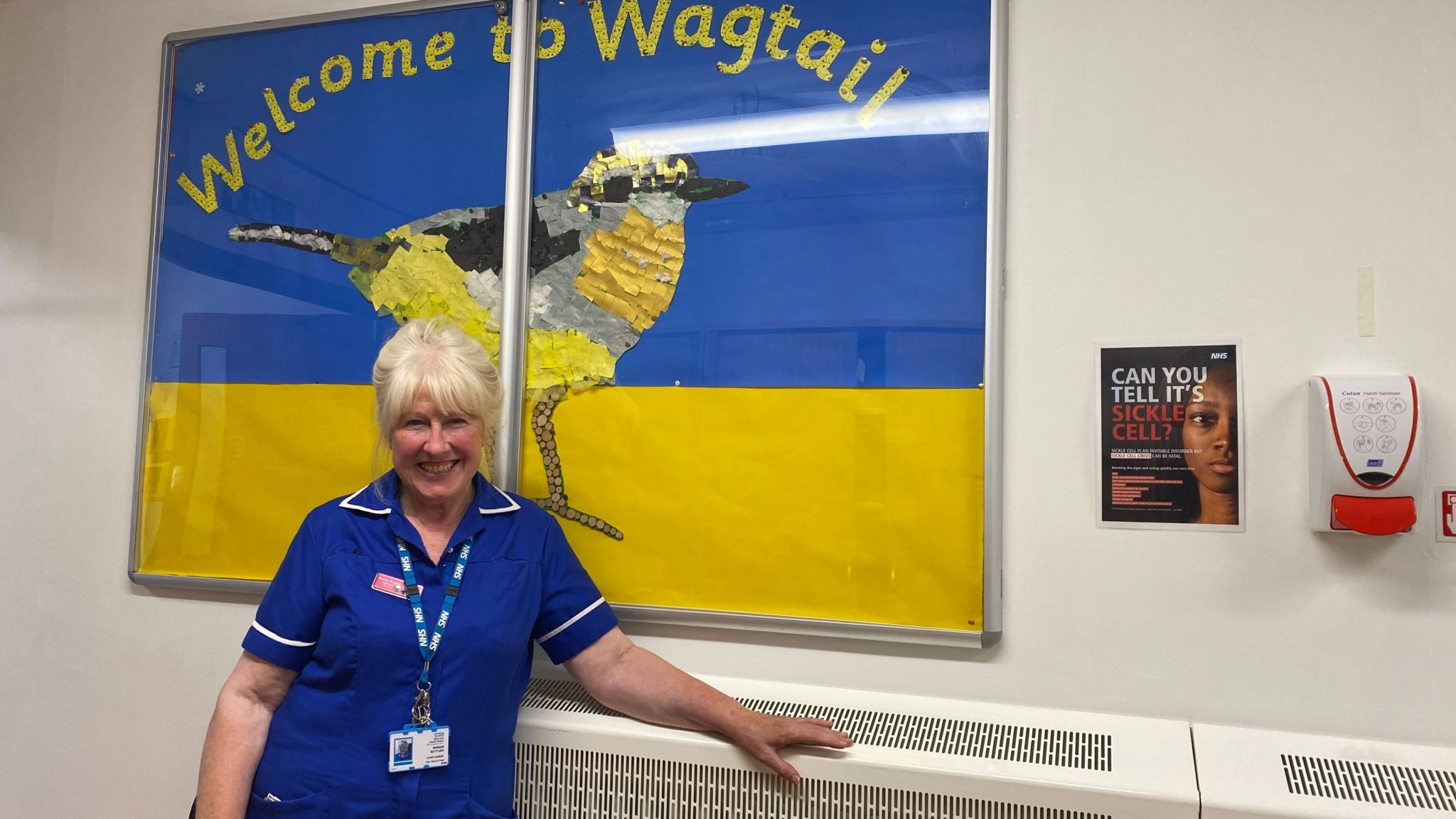 Marian Bottjer smiles at the camera, wearing her blue nursing uniform, as she leans against a radiator underneath a painting of a gold bird in the hospital corridor. 