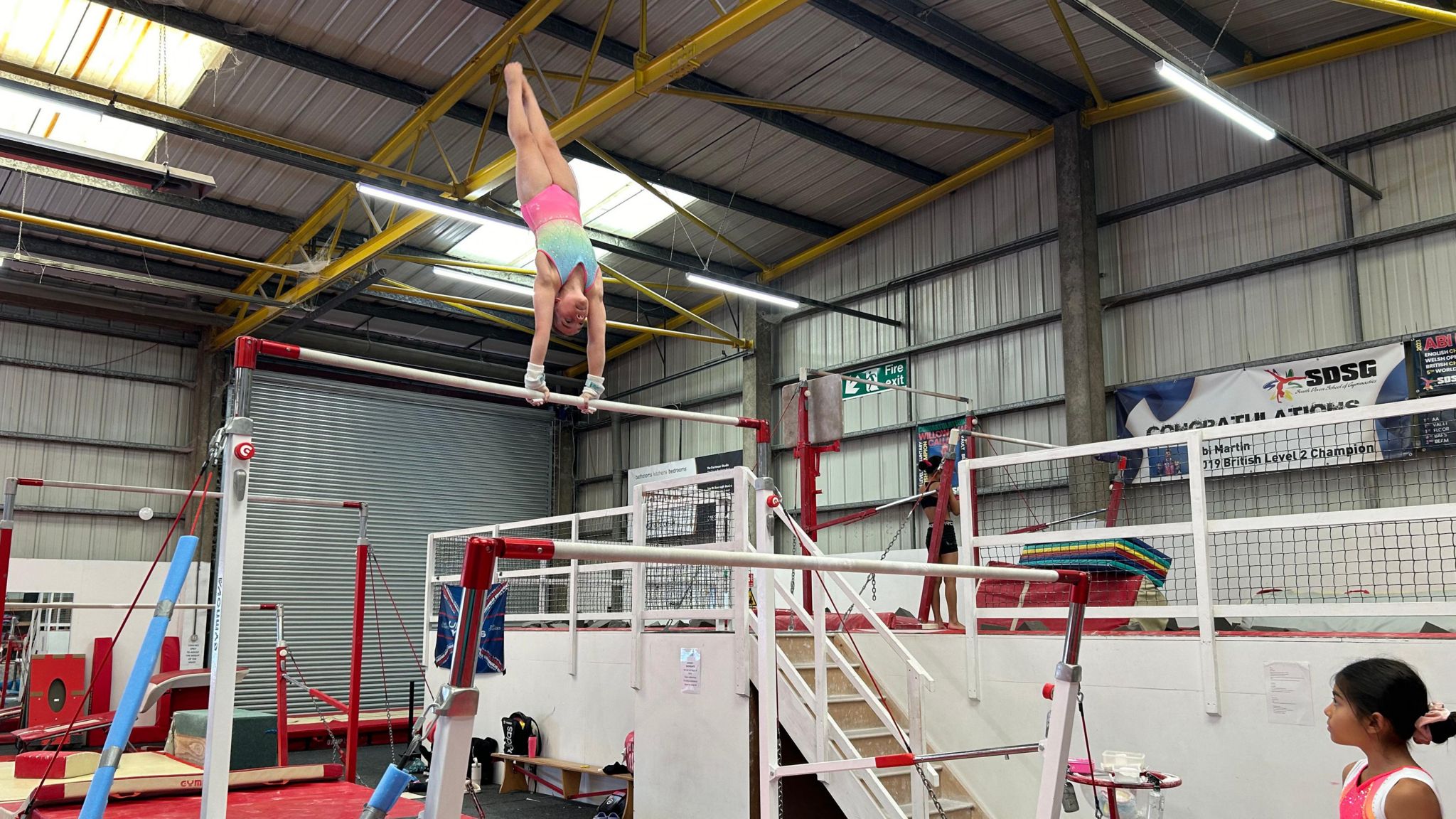 A young gymnast in a blue, yellow and pink leotard performing a move called a giant which sees her doing a spinning handstand on the uneven bars while a team-mate watches on