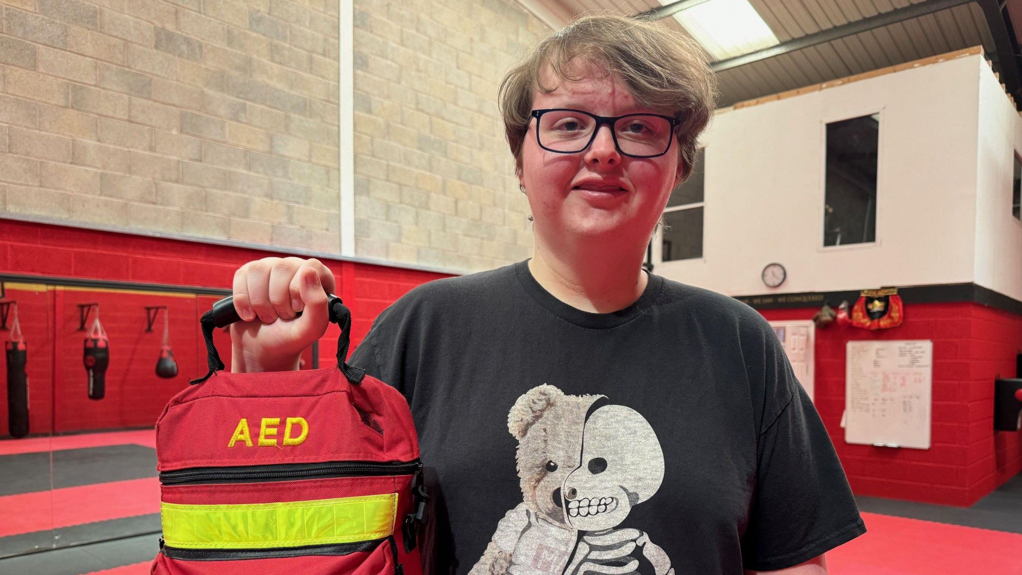 A teenage boy in a black tshirt holds up a red defibrillator in a room with gym equipment and matting in the background