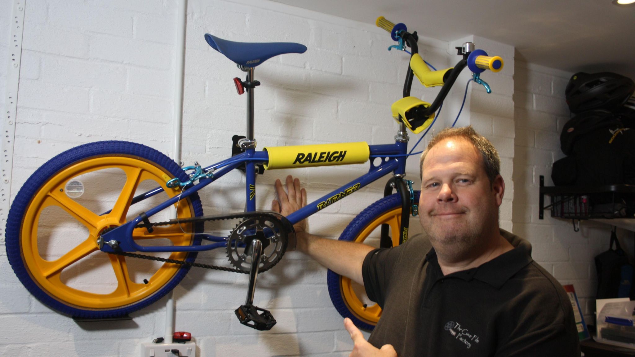 Simon Papouis wearing a black shirt and smiling at the camera with his right hand pressed against a white brick wall. Hanging on the wall is a blue and yellow bike.