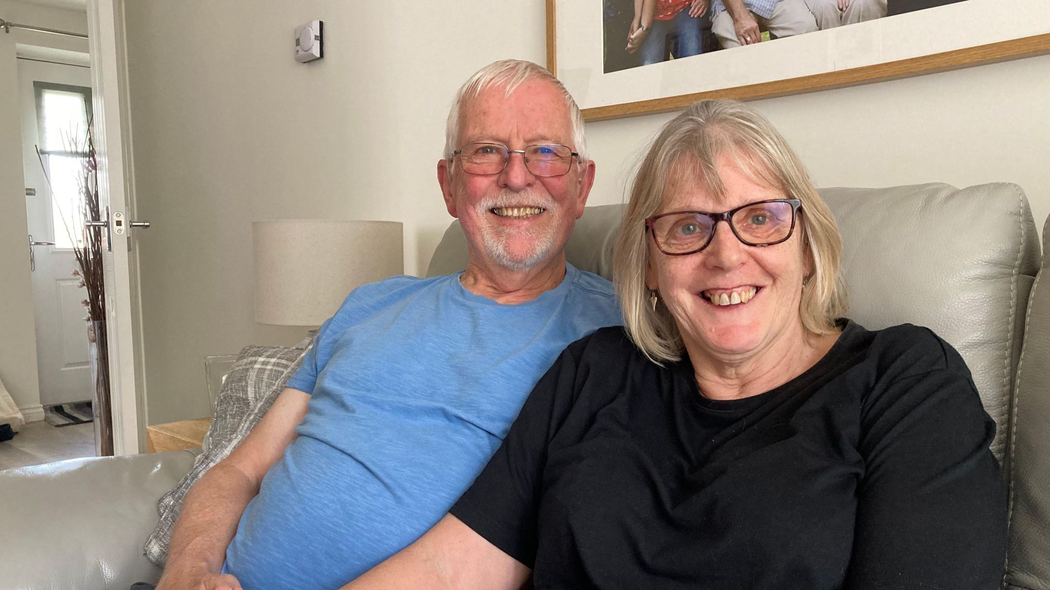 A man in a blue T-shirt and a woman in a black T-shirt sit on a light coloured sofa with a open door in the background