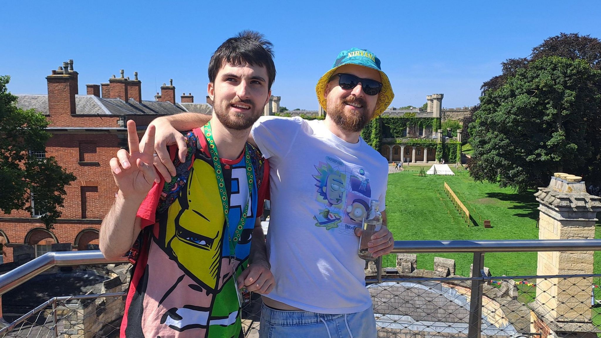 Two men standing arm in arm, the one on the lefts is wearing a colourful marvel cartoon patterns tshirt and the man on the right is wearing a white shirt with cartoons on the chest and he is also wearing a hat