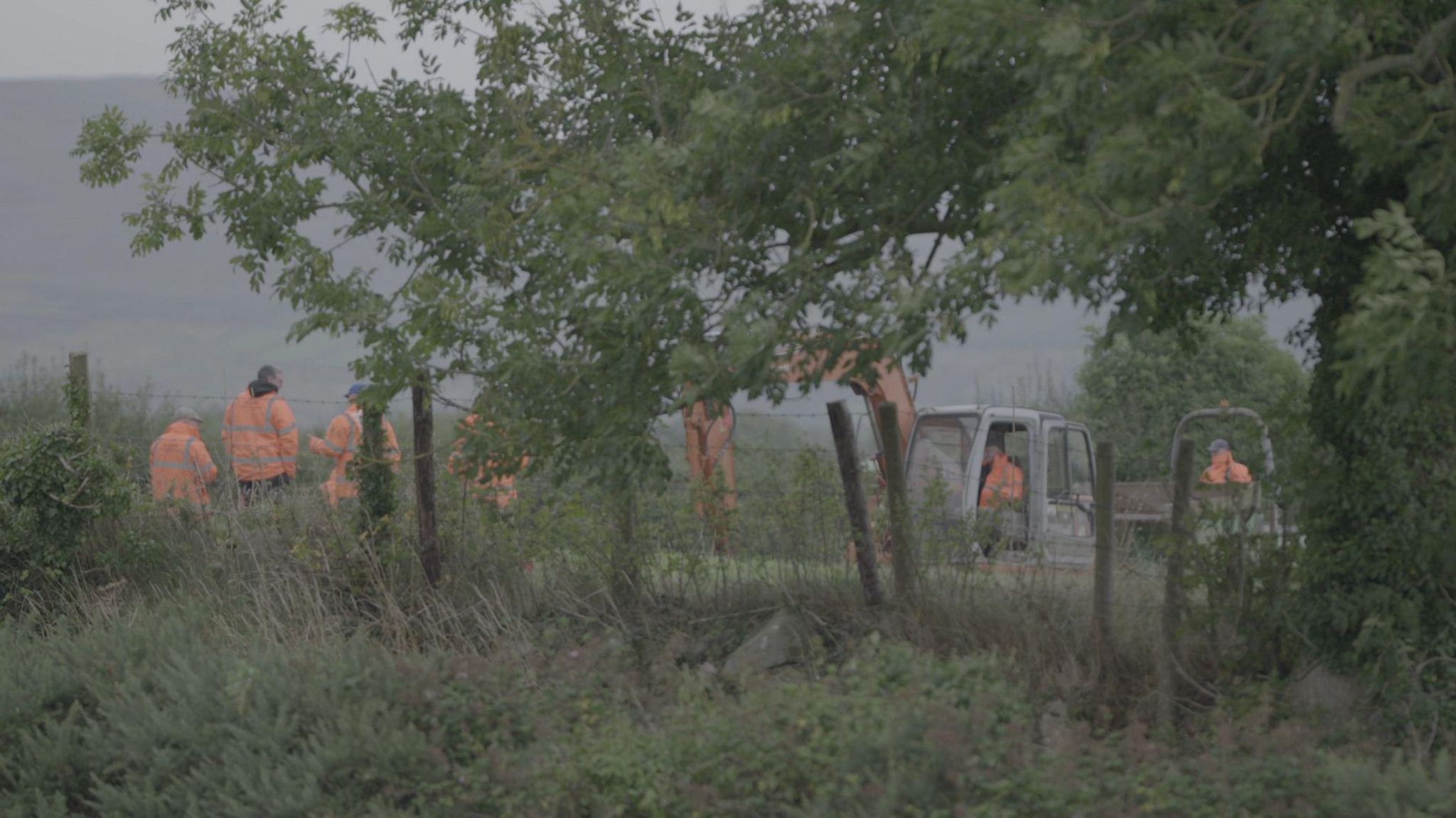 People in hi-vis jackets in a field. There is a digger in the field also. The photo is taken through trees.