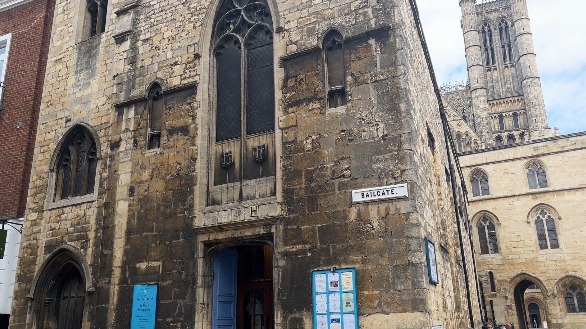 St Mary Magdalene Church is a stone building with a blue front door and noticeboard. Arched windows and a street sign reading bailgate lincoln