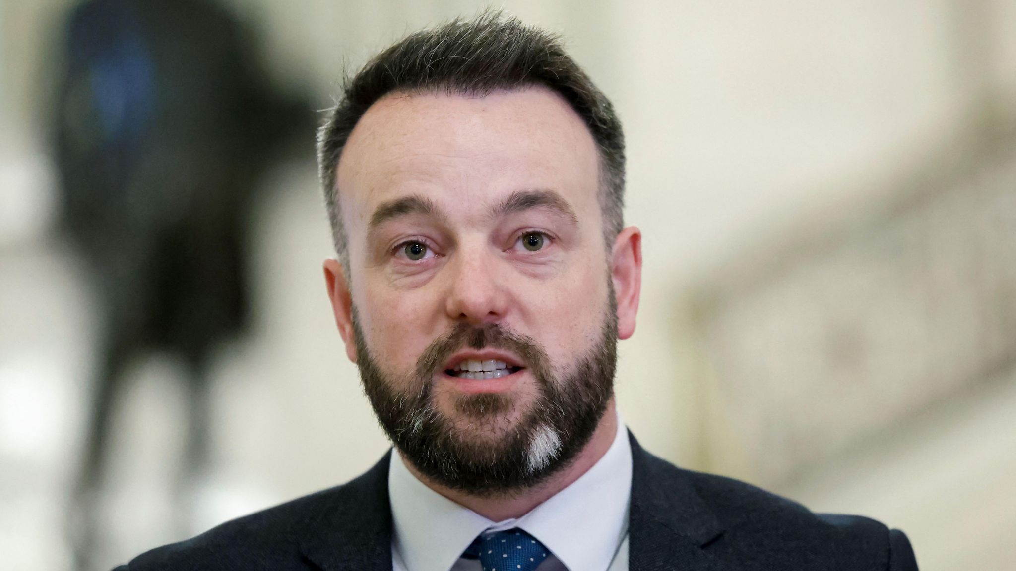 Colum Eastwood - a man with dark hair and beard wearing a suit looks beyond the camera