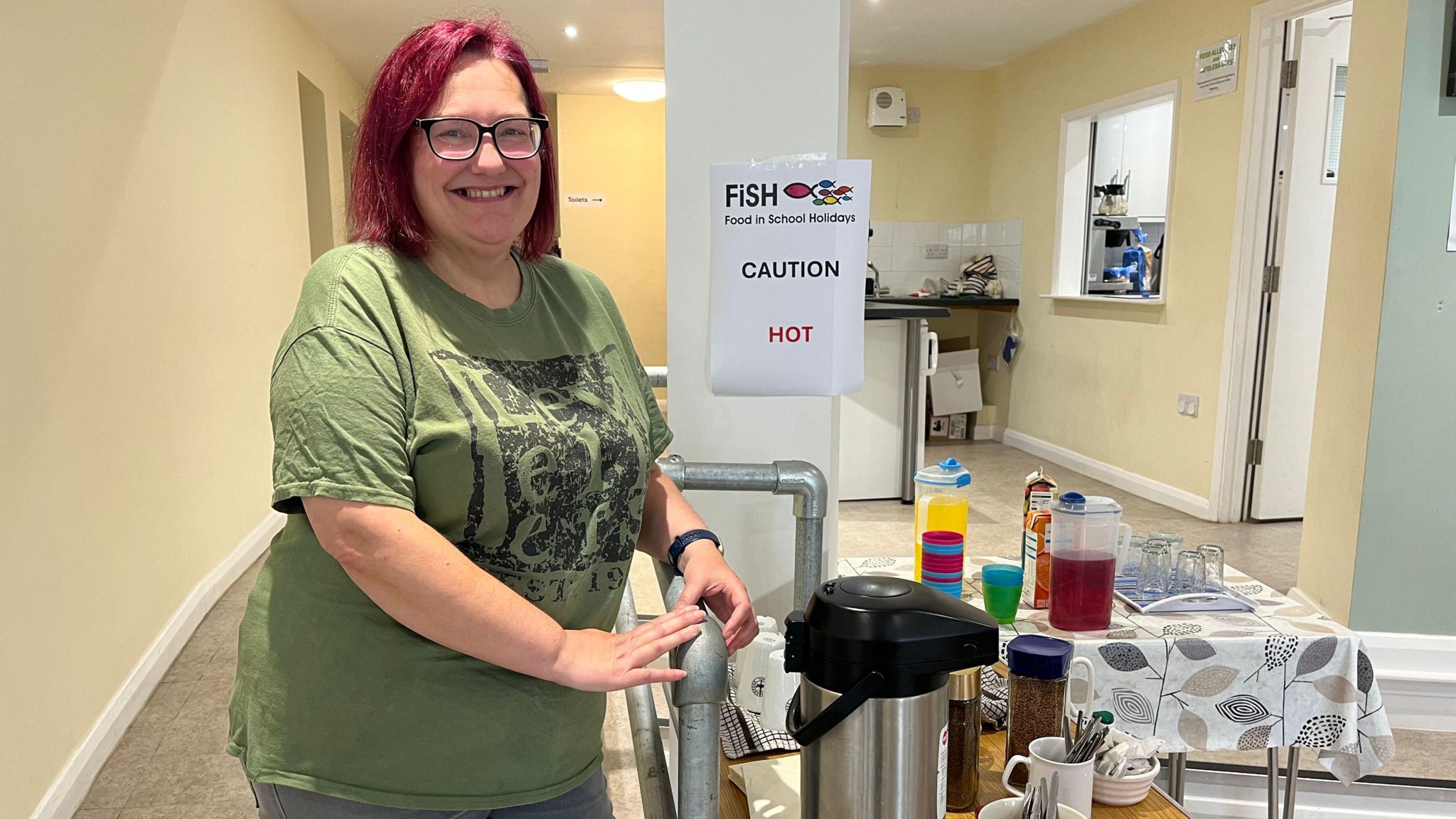 Reverend June Love stands next to a table filled with hot and cold drinks on offer at the buffet. She has bright red hair wears glasses and a broad smile. She is wearing an olive T-shirt