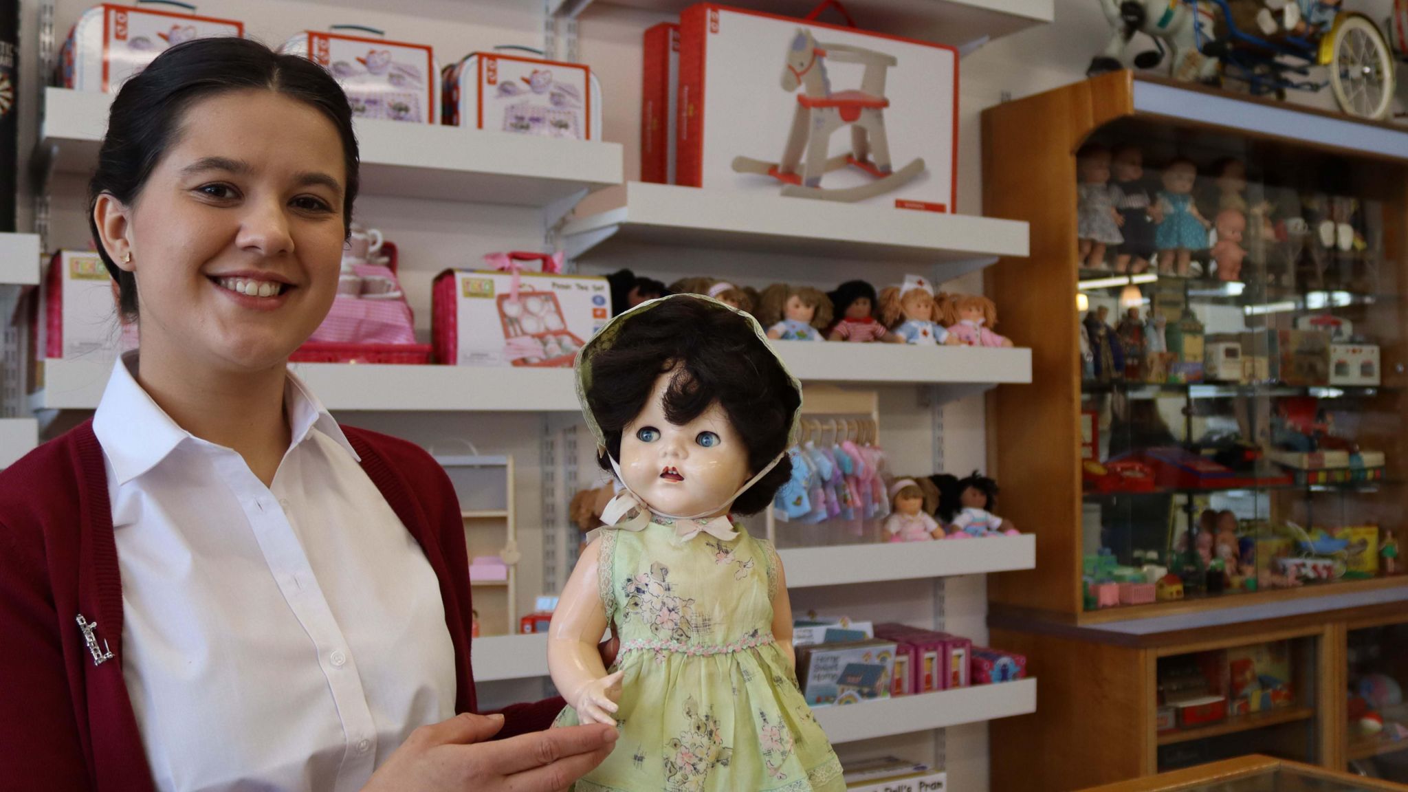 A woman in a red cardigan holds a vintage doll in a yellow dress in front of toy displays