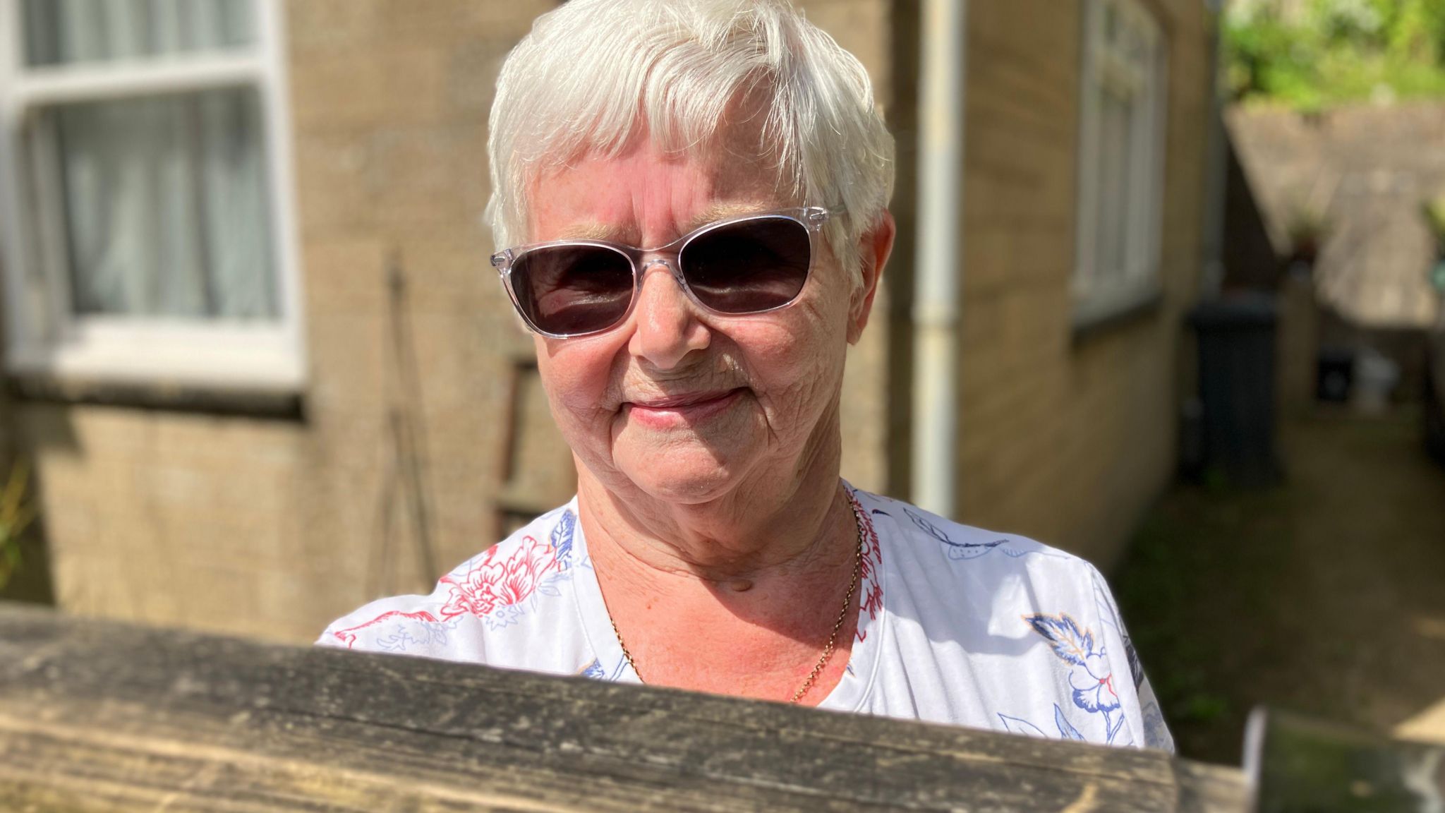 Ann Townend smiling in front of her home, wearing sunglasses and white top.