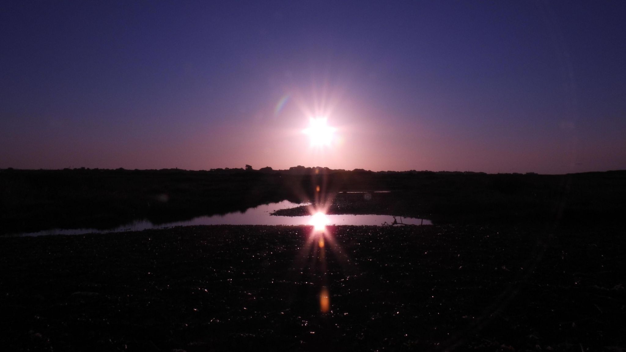 The sun is reflected in a pool of water. The sky is a beautiful purple colour while the rest of the picture is in darkness.