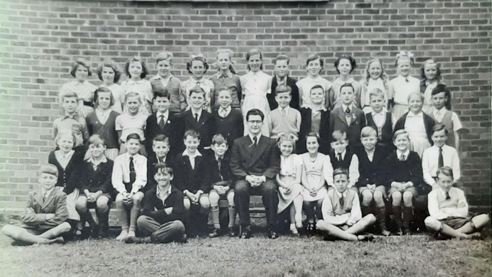 A black and white school photo showing a male teacher in a suit and tie surrounded by his young students.