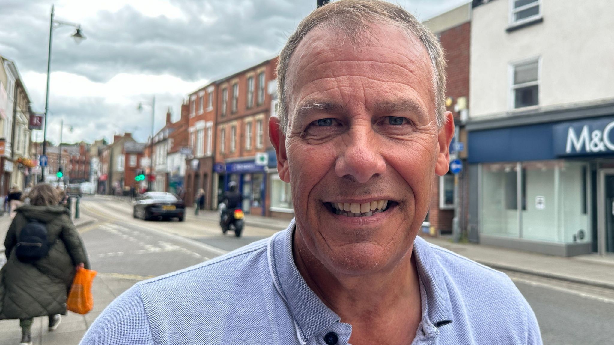 Paul Sanders, wearing an open collar blue shirt and posing for a picture in the street. Shoppers and vehicles can be seen in the background