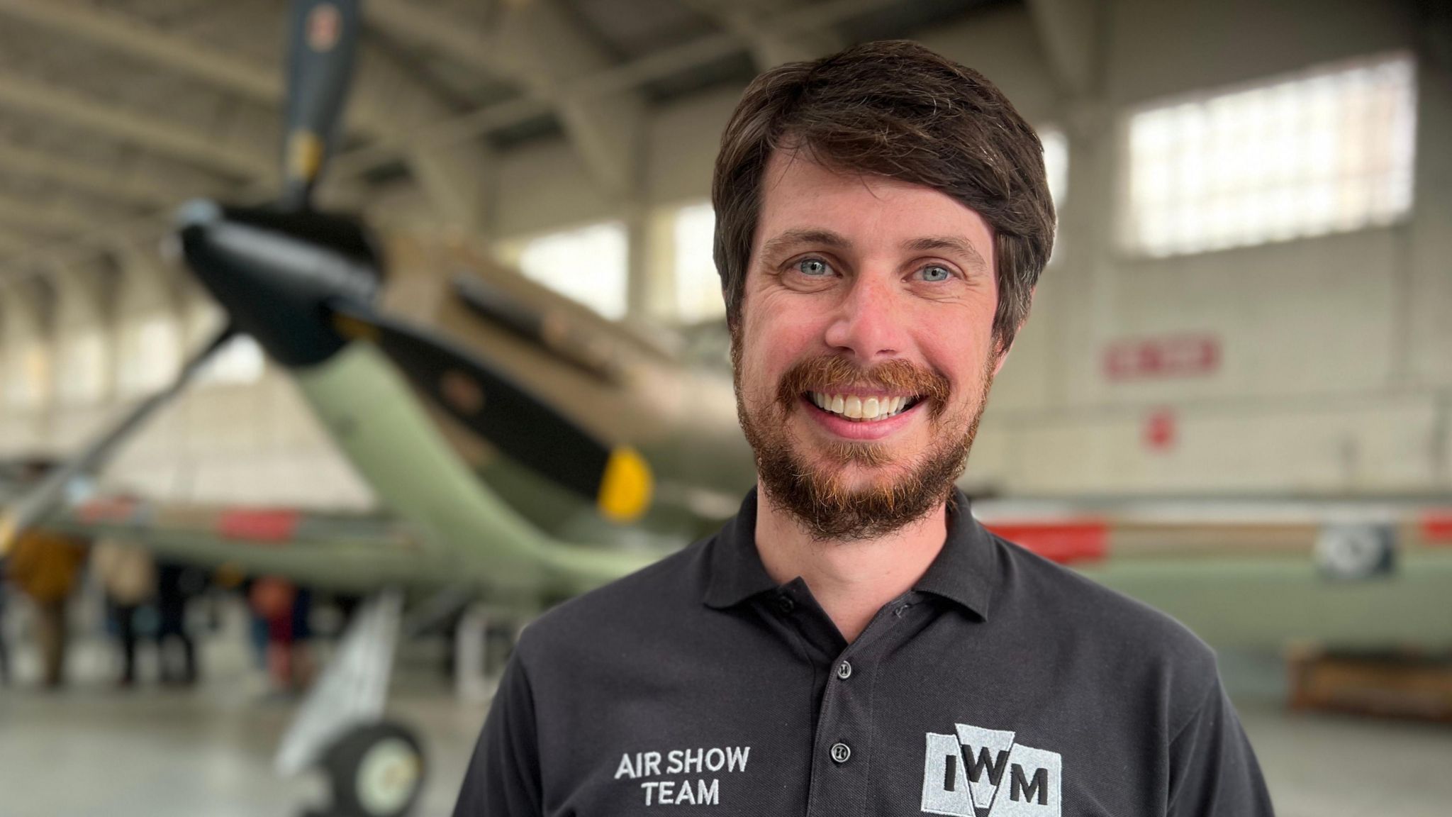 Phil Hood wearing a shirt saying Air Show Team and IWM in a museum hangar with an aircraft behind him