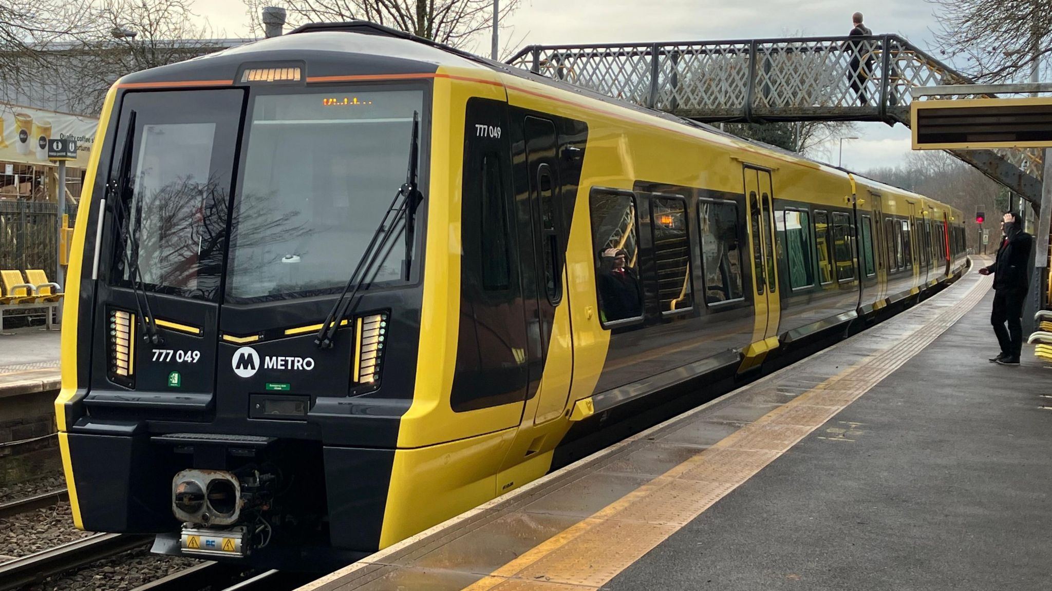 A yellow and black Metro train with three carriages stands at a platform, with a pedestrian bridge spanning the tracks in the background