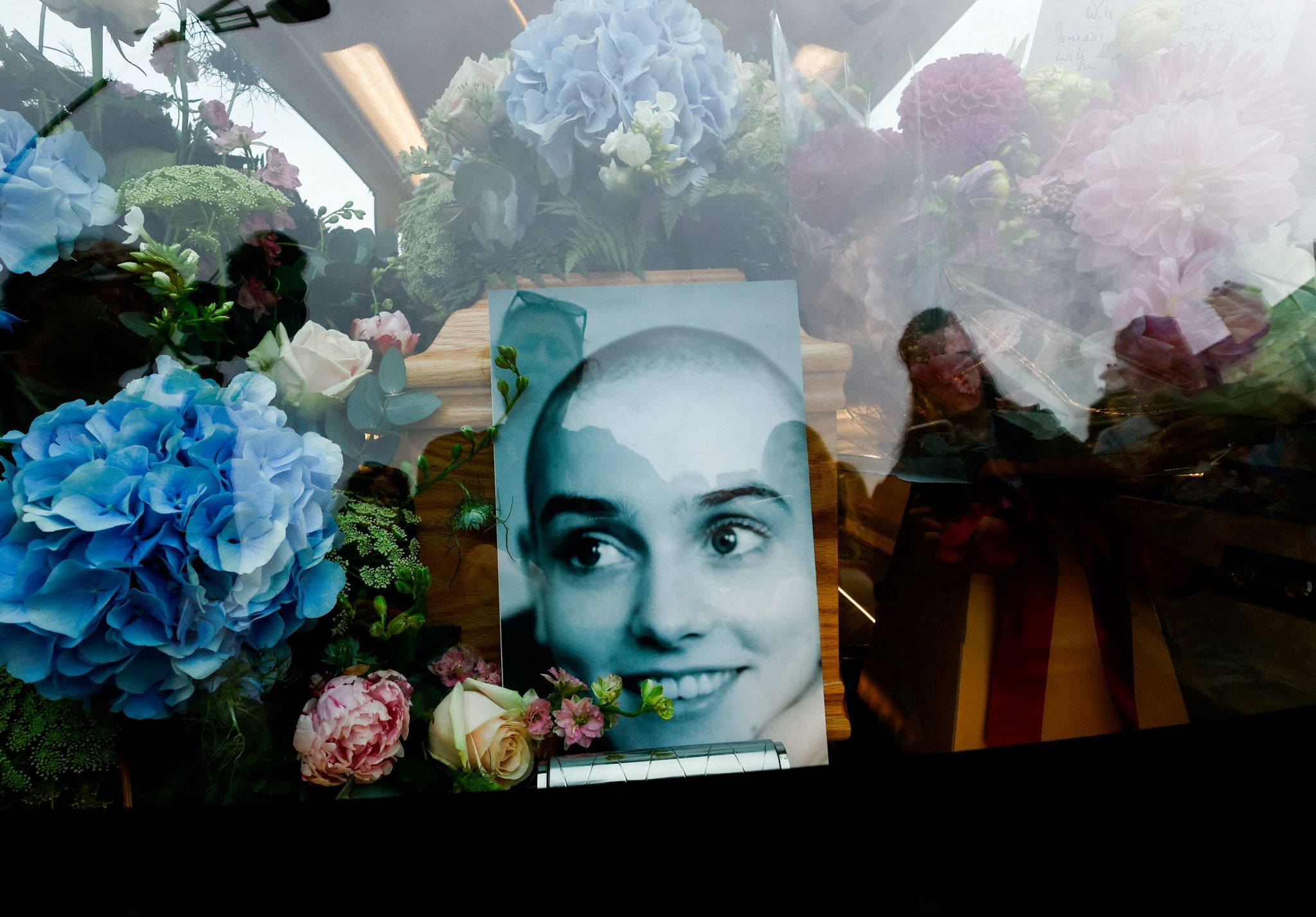 A black and white photo of a young Sinead O'Connor sits in front of her coffin, surrounded by flowers, inside a hearse