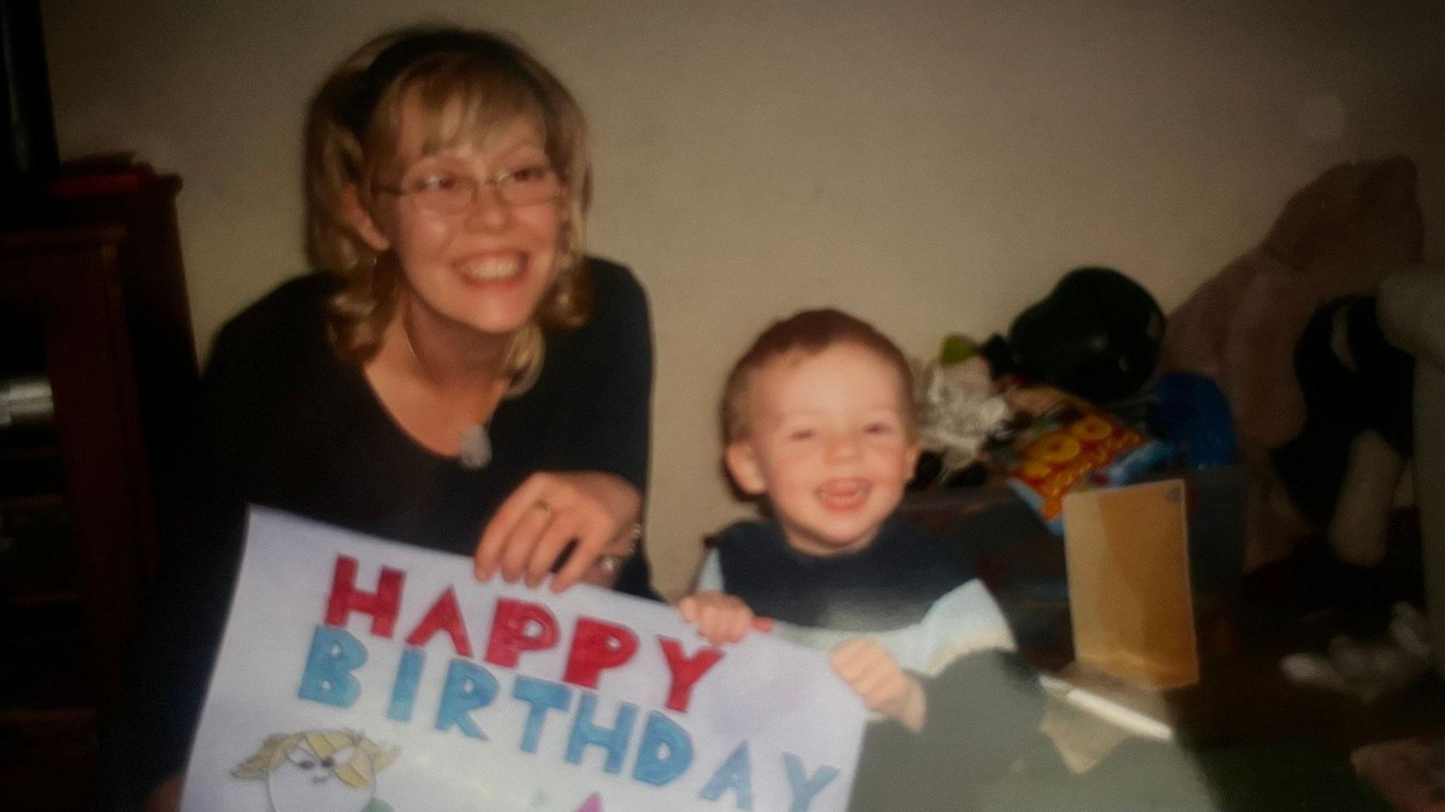 A young Alfie Watts and his mother both smile as they both hold a big piece of paper bearing the words "happy birthday"