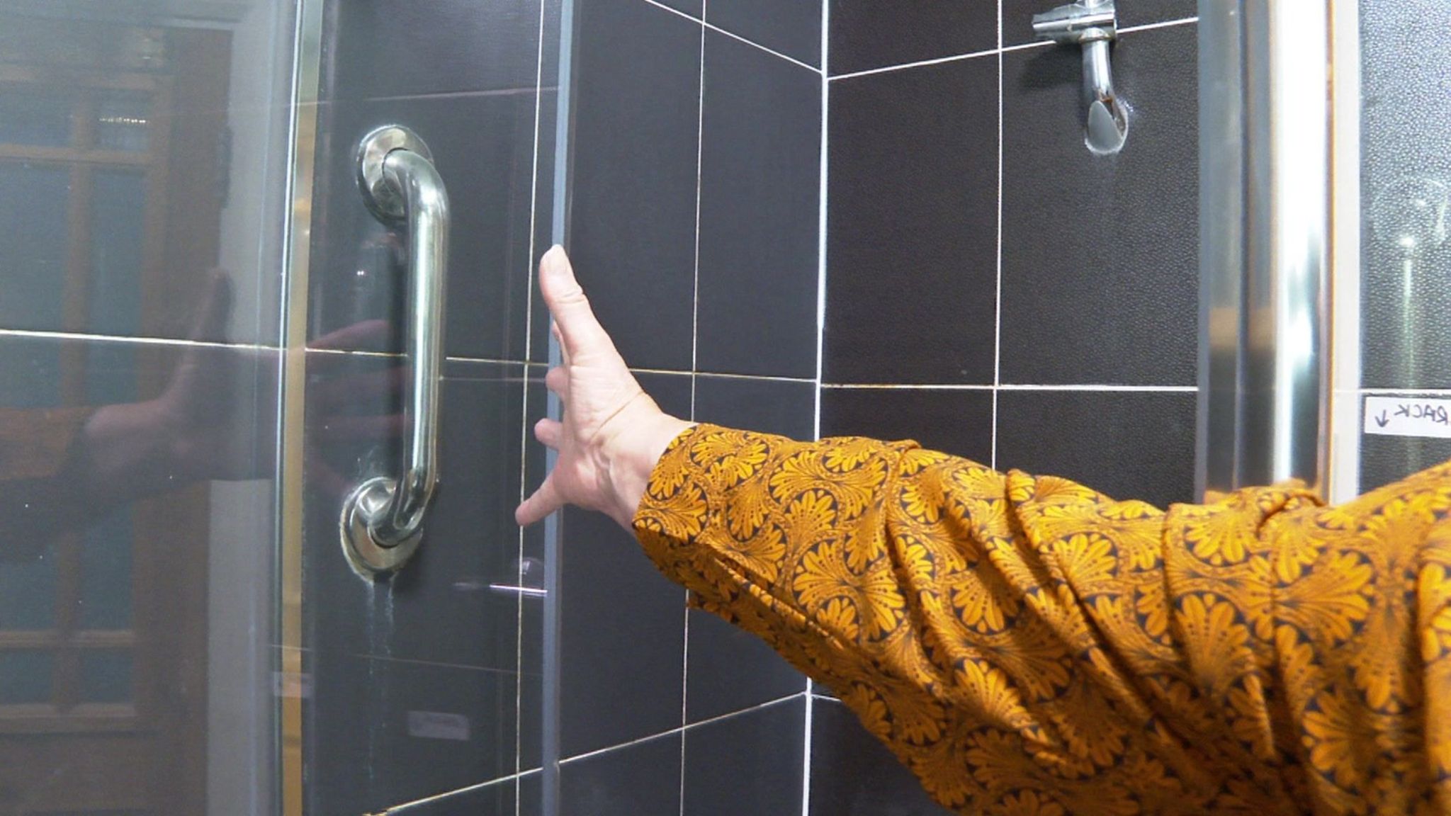 A woman's arm is unable to reach a grab handle blocked by a glass shower door