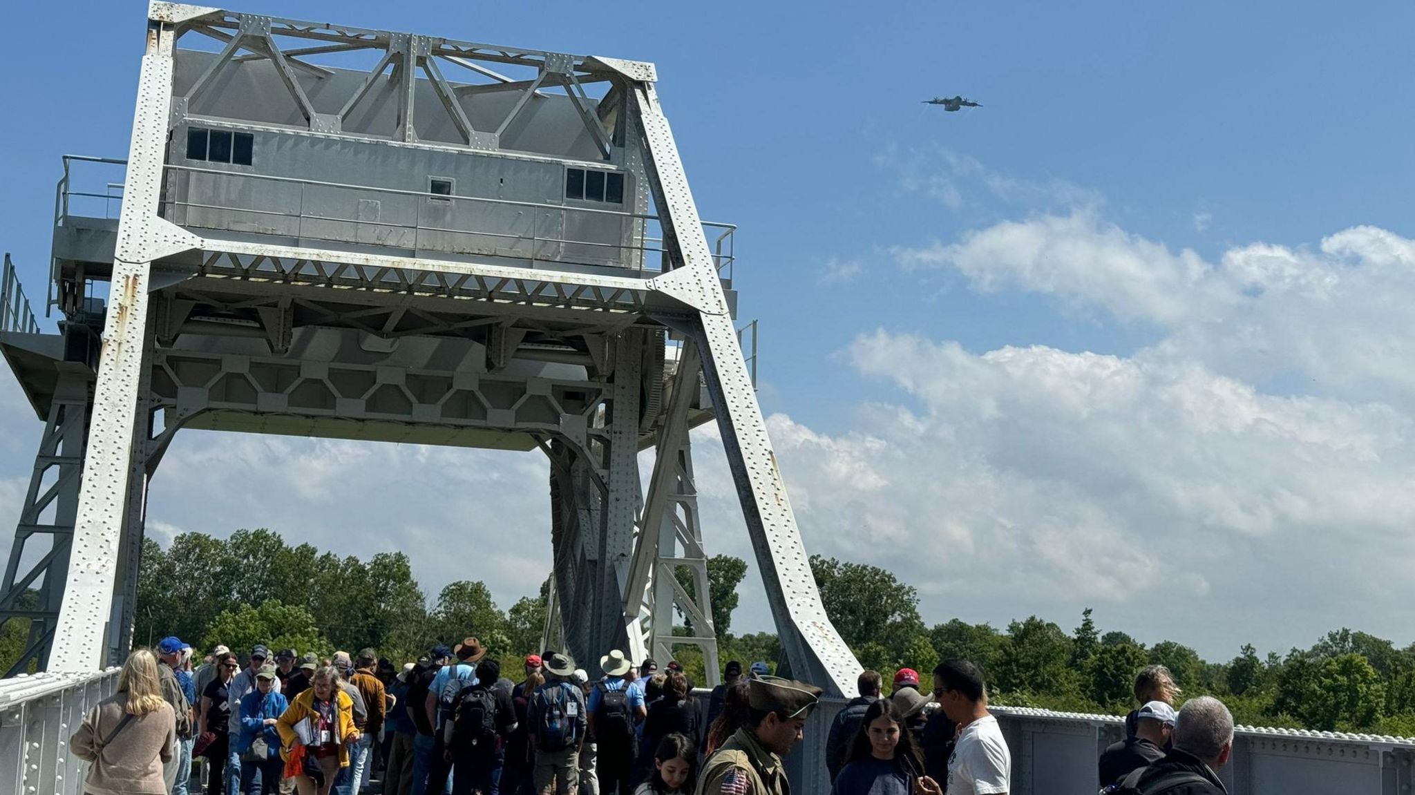 A military aircraft in the distance flying past a bridge full of people