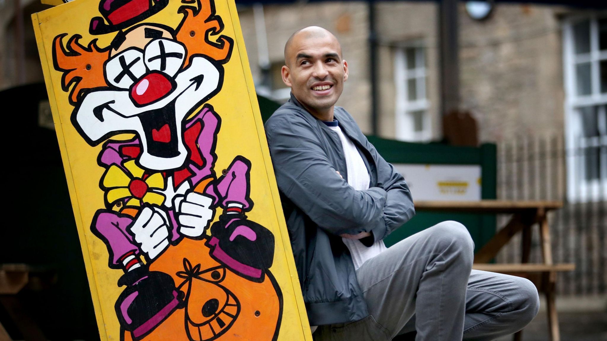 Comedian Masai Graham wears a grey jacket and jeans and poses in front of a colourful sign with a clown riding a space hopper painted on it.