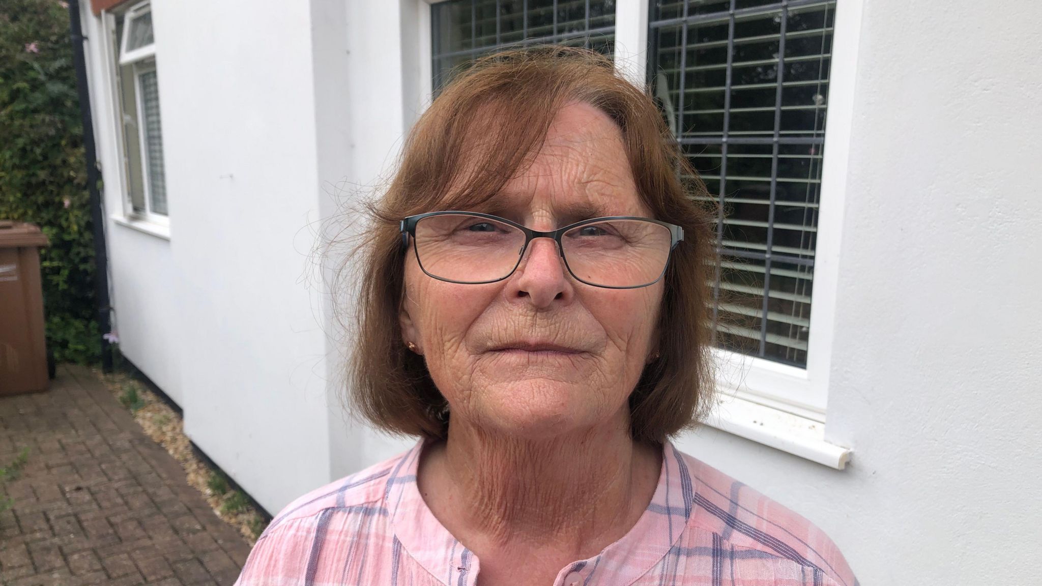 Rita Phillips looks unhappy as she gazes directly at the camera. She is photographed outside a white house, wearing a pink blouse and glasses. 