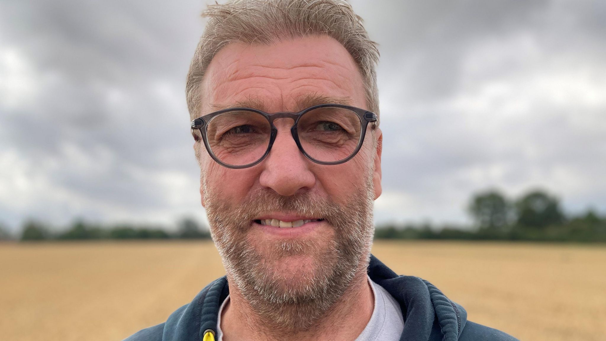 A head and shoulders photograph of farmer Carl Benson in a field with trees in the distance. He has light brown hair, black circle-rimmed glasses and a light brown beard.